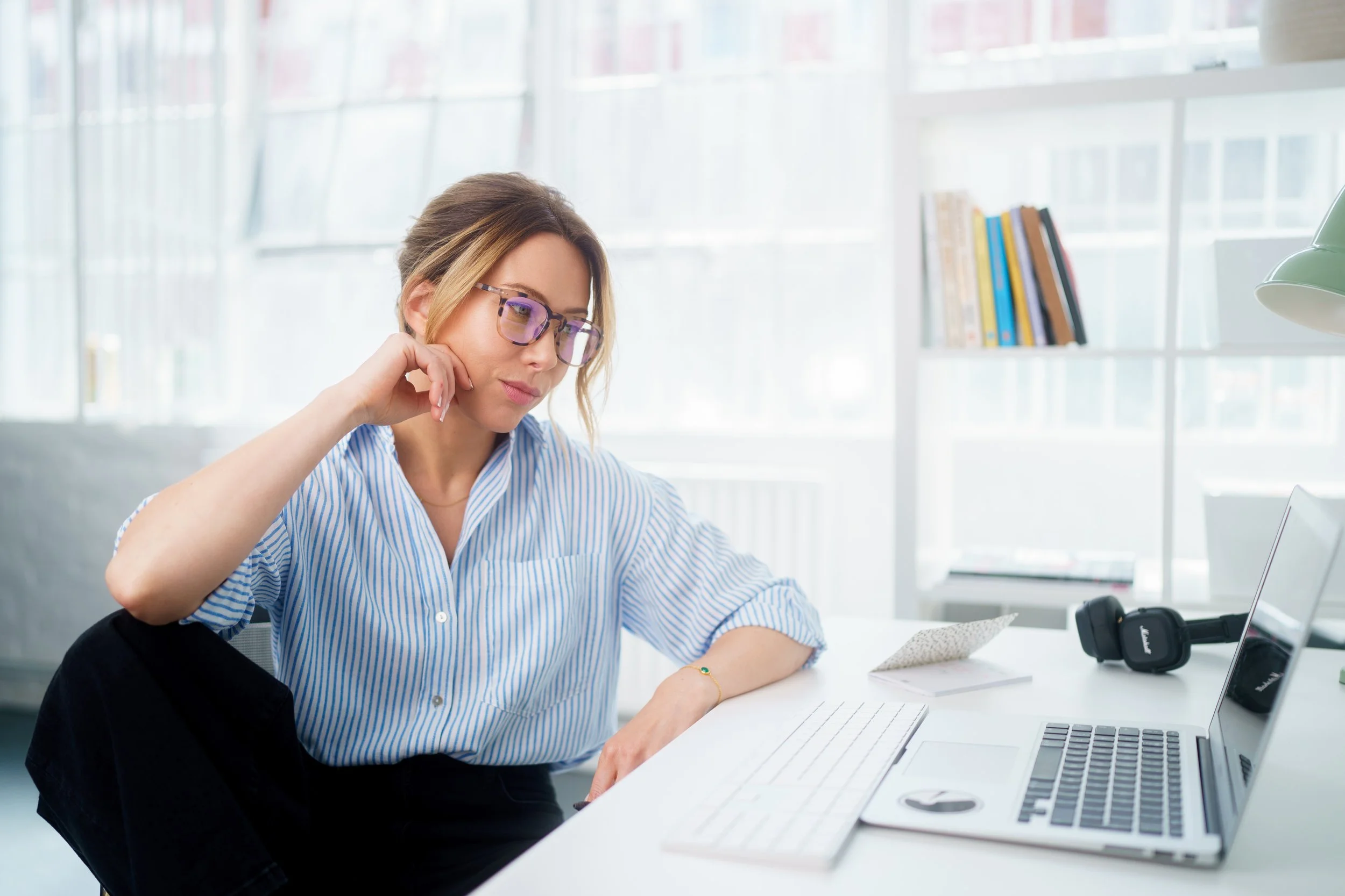 A woman with glasses and a woman with dark hair are working together at a white desk in a bright office space with large windows. The woman with glasses appears focused and is resting her head on her hand, while the other woman is writing or taking notes.