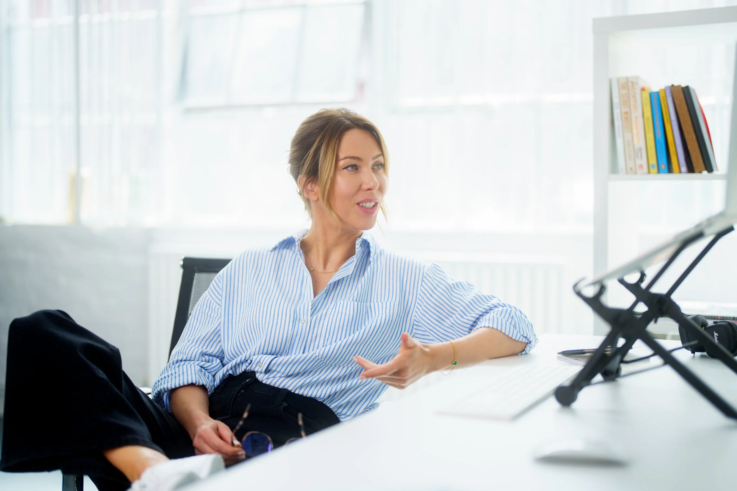 A woman with blonde hair in a loose bun, wearing a blue and white striped shirt, sitting at a white table in a bright, modern office space. She appears to be talking and gesturing with her right hand. There are books on a white shelf behind her and a stroller in front of her on the table.