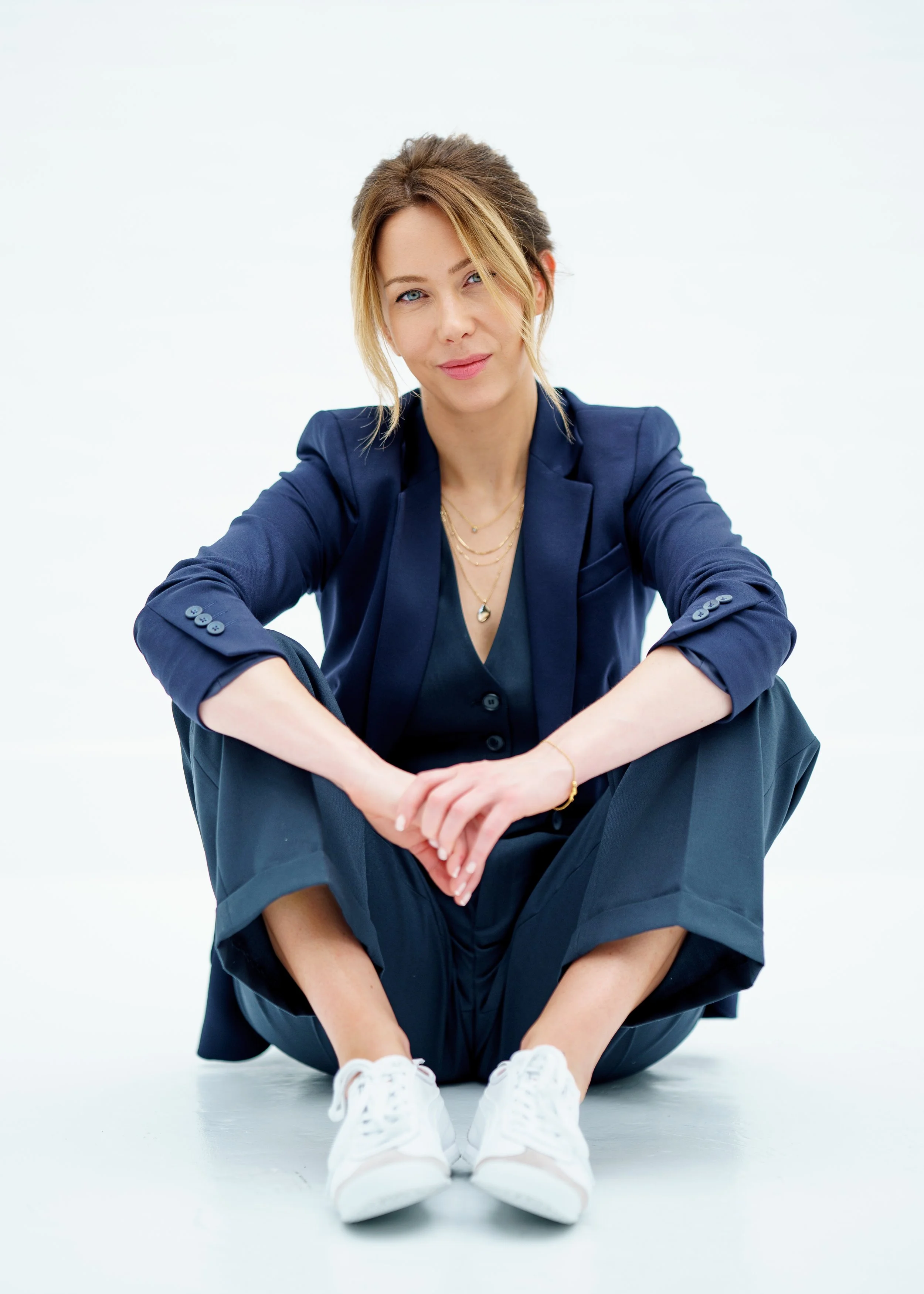 Woman sitting on the floor with legs crossed, wearing a navy blazer and pants, white sneakers, and layered necklaces, looking at the camera with a slight smile against a white background.