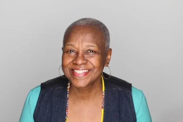 Close-up of a smiling older woman with short gray hair, wearing hoop earrings, a turquoise shirt, and a black vest, against a plain light gray background.