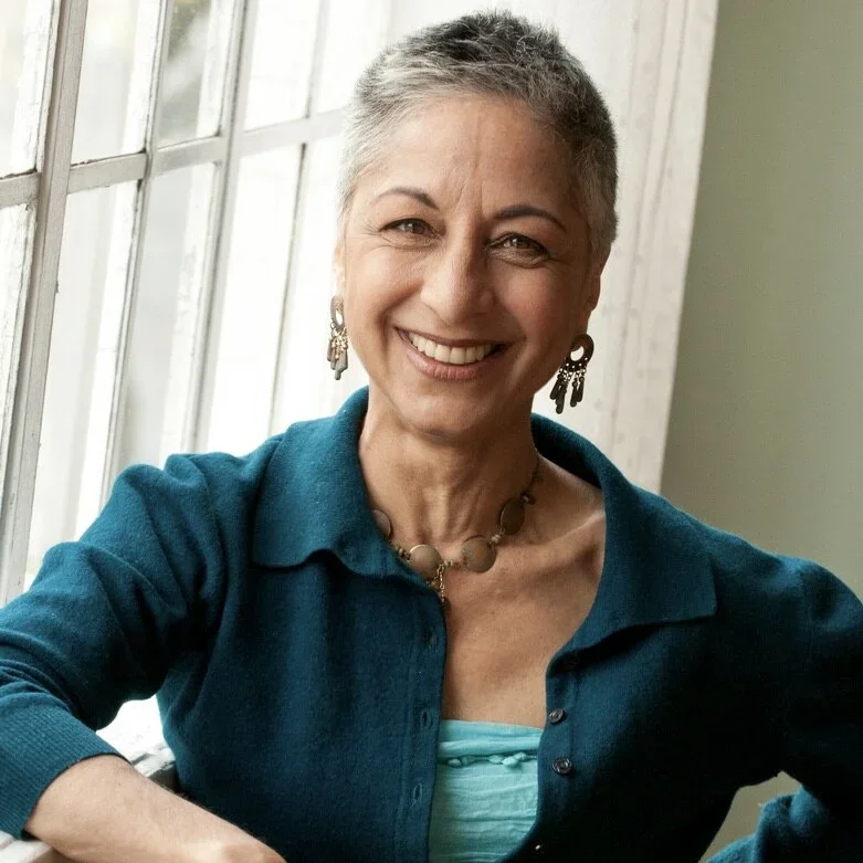 A smiling woman with short gray hair wearing earrings, a beaded necklace, a blue cardigan, and a teal top, sitting by a window.