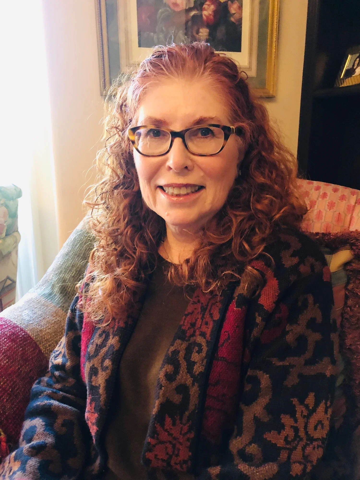 A woman with curly red hair and glasses sitting indoors, smiling at the camera. She is wearing a black top and a colorful patterned cardigan. Background includes a framed painting or photograph and a black bookshelf.