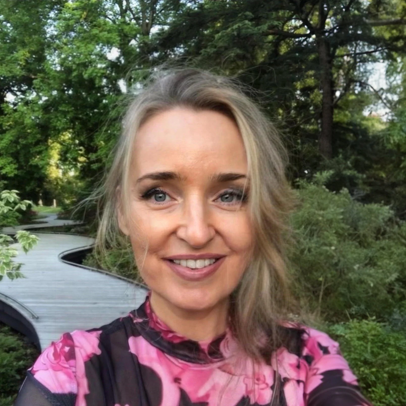 A woman with blonde hair and blue eyes smiling outdoors in front of green trees, wearing a pink and purple patterned shirt.