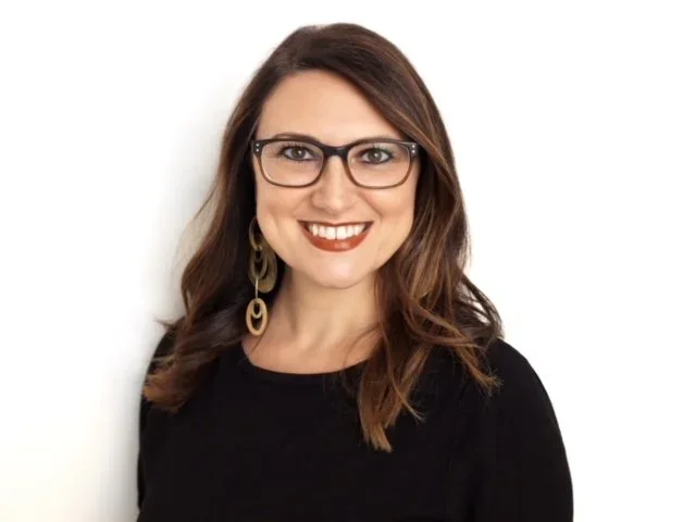A woman with long brown hair, glasses, and earrings, smiling against a white background.