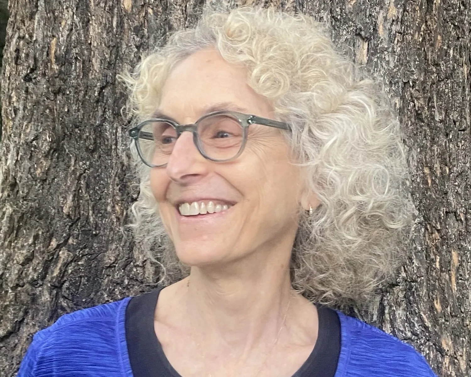 A woman with curly gray hair and glasses smiling in front of a large tree trunk, wearing a blue shirt.