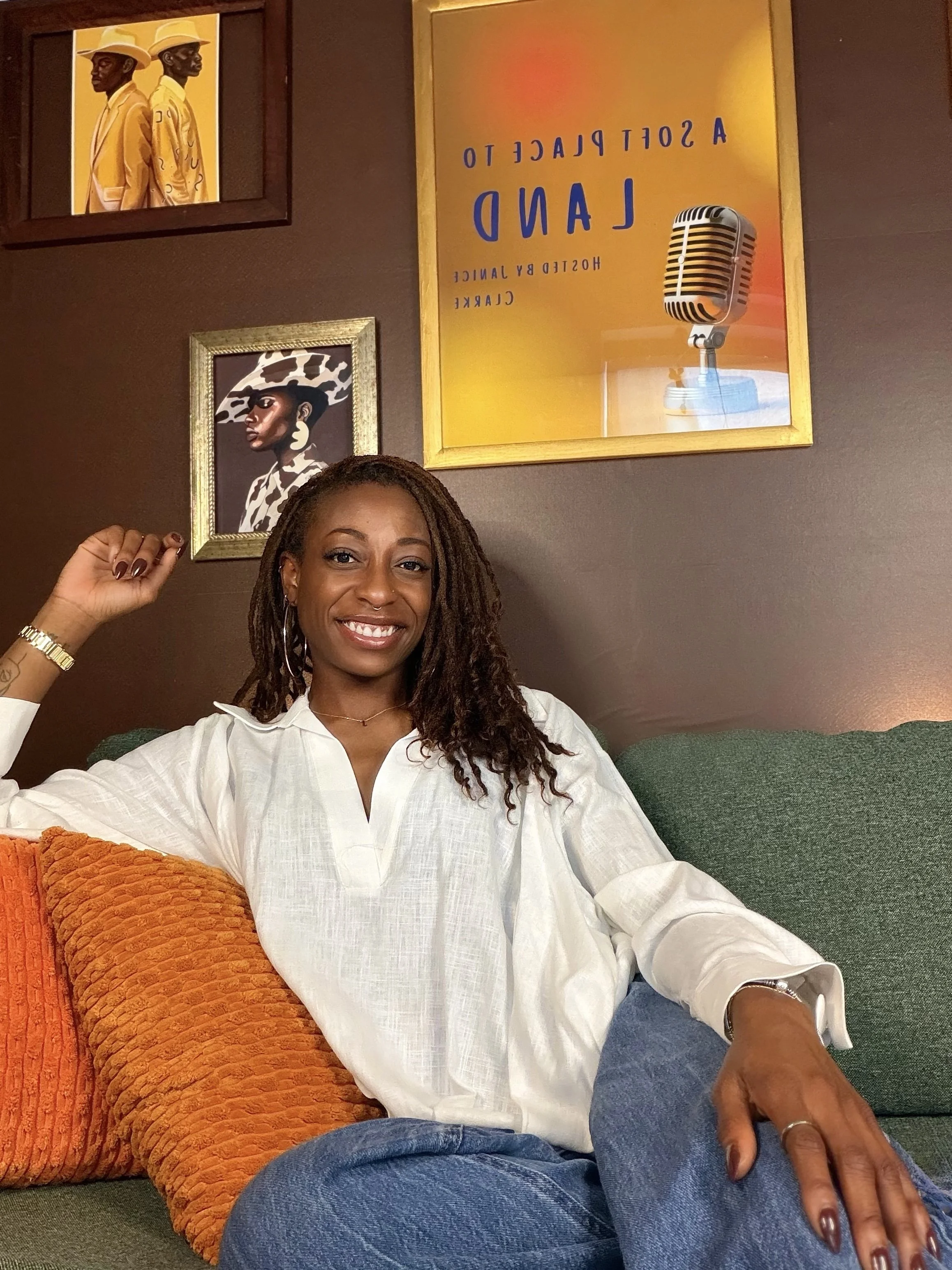 A woman with dreadlocks and jewelry is sitting on a green couch, smiling at the camera. She is wearing a white shirt and blue jeans. There are framed pictures hanging on the brown wall behind her, including one of two women in yellow suits and hats, and another of a woman in a cow-printed outfit and hat. There is also a framed poster with text and a vintage microphone illustration.
