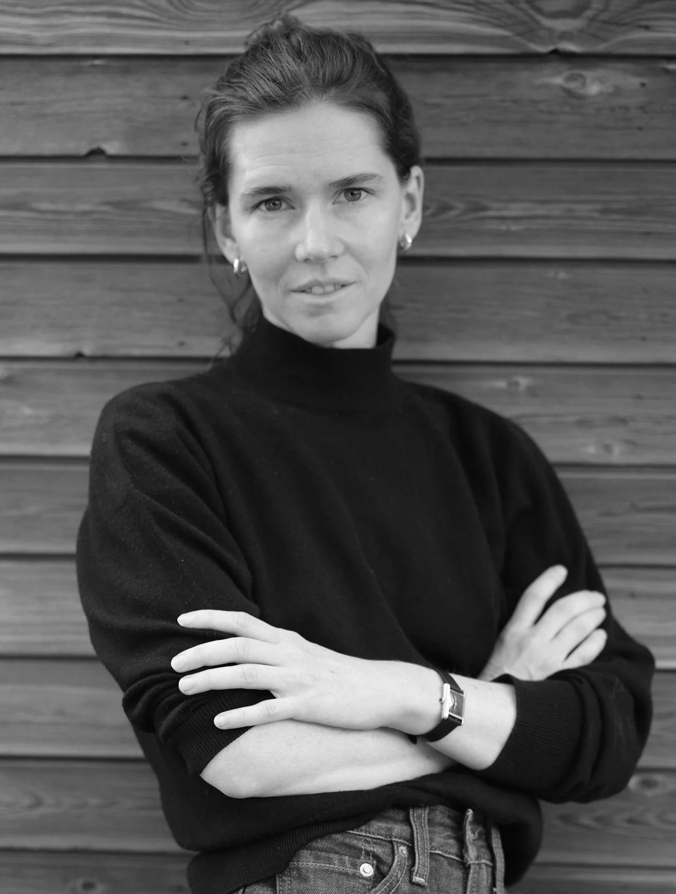 Black-and-white portrait of a woman in a black turtleneck with crossed arms, standing against a wooden plank wall.