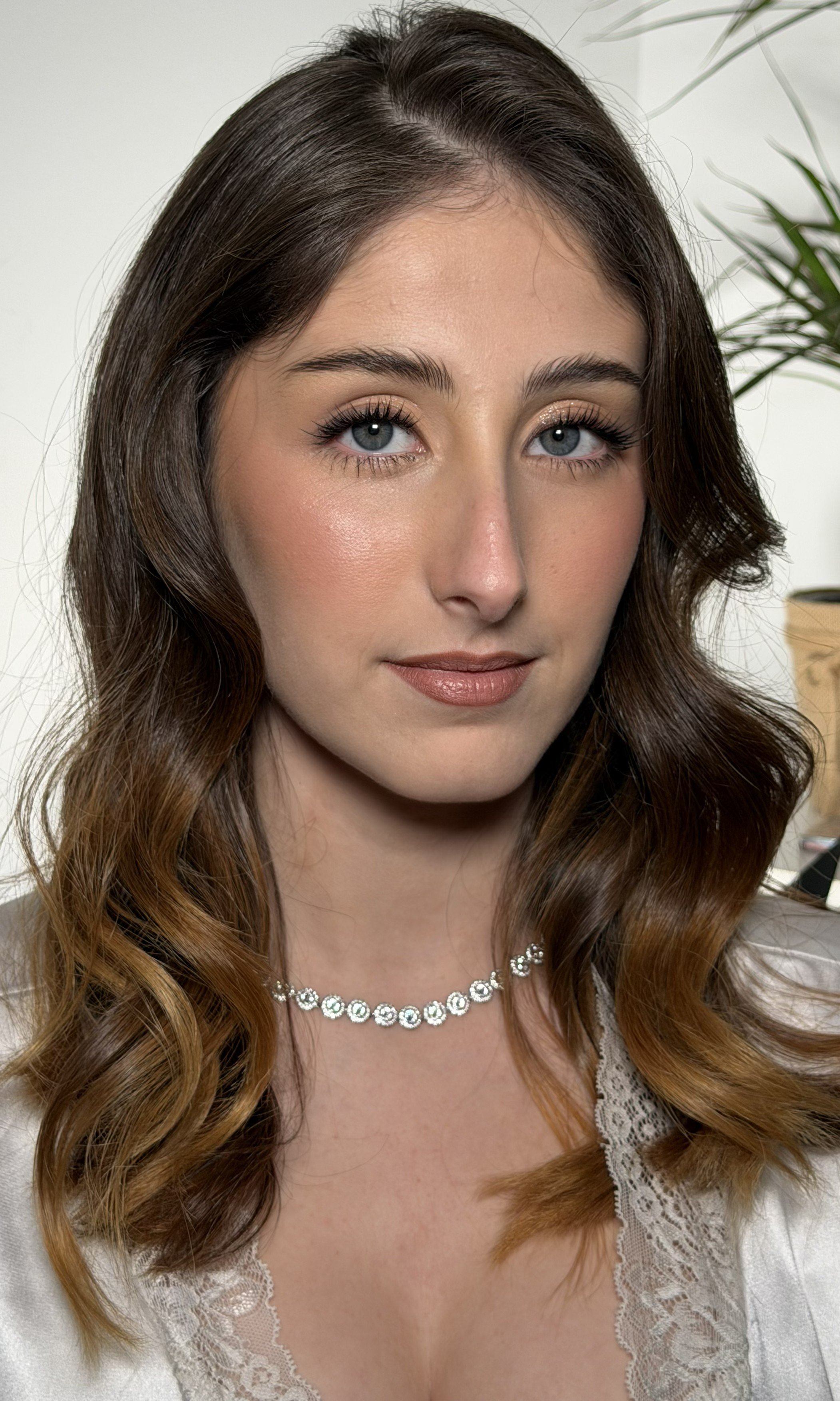 Close-up of a woman with wavy, brown hair and blue eyes, wearing a pearl necklace and lace-trimmed top, smiling softly indoors.