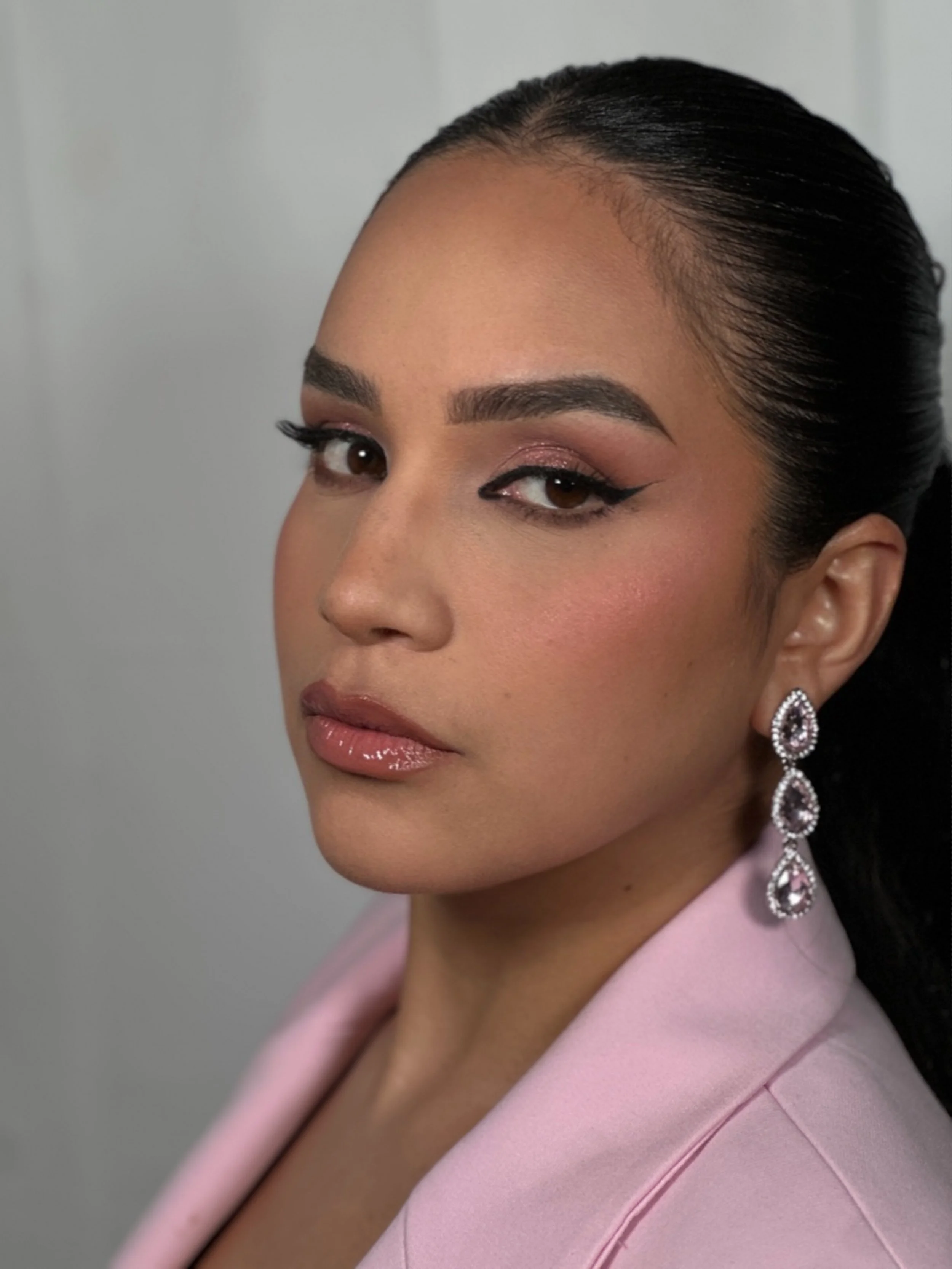 Close-up of a woman with dark hair sleeked back, wearing makeup with winged eyeliner, pinkish eyeshadow, and glossy lips, accessorized with large sparkling teardrop earrings, dressed in a pink blazer, against a neutral background.