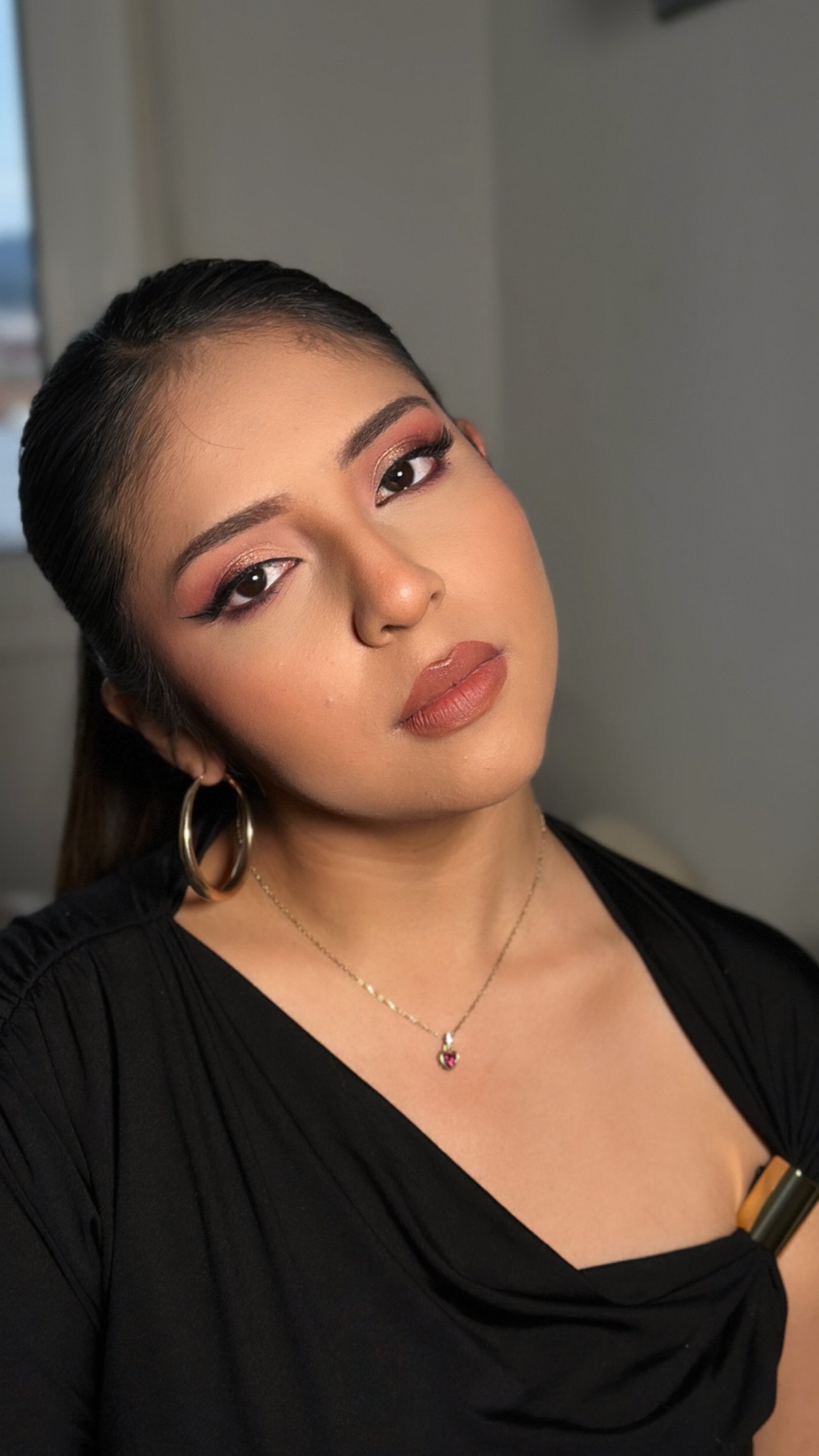 A young woman with dark hair, makeup, and hoop earrings is posing for a photo, wearing a black top and a necklace with a pendant.