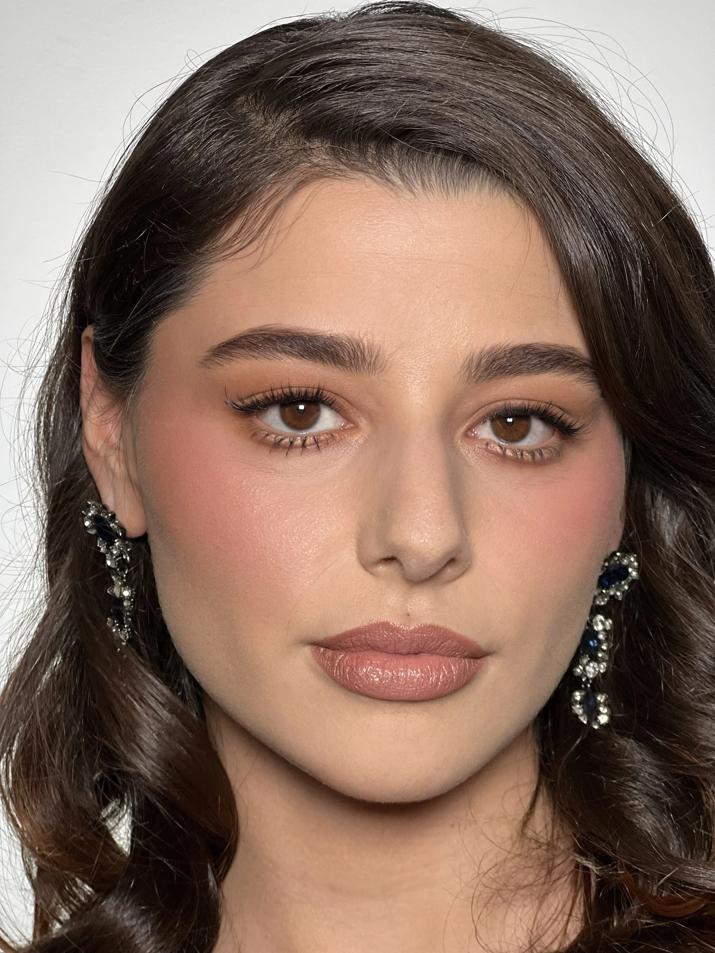 Close-up of a woman's face focusing on her eyes, lips, and earrings, with dark brown, wavy hair and makeup.