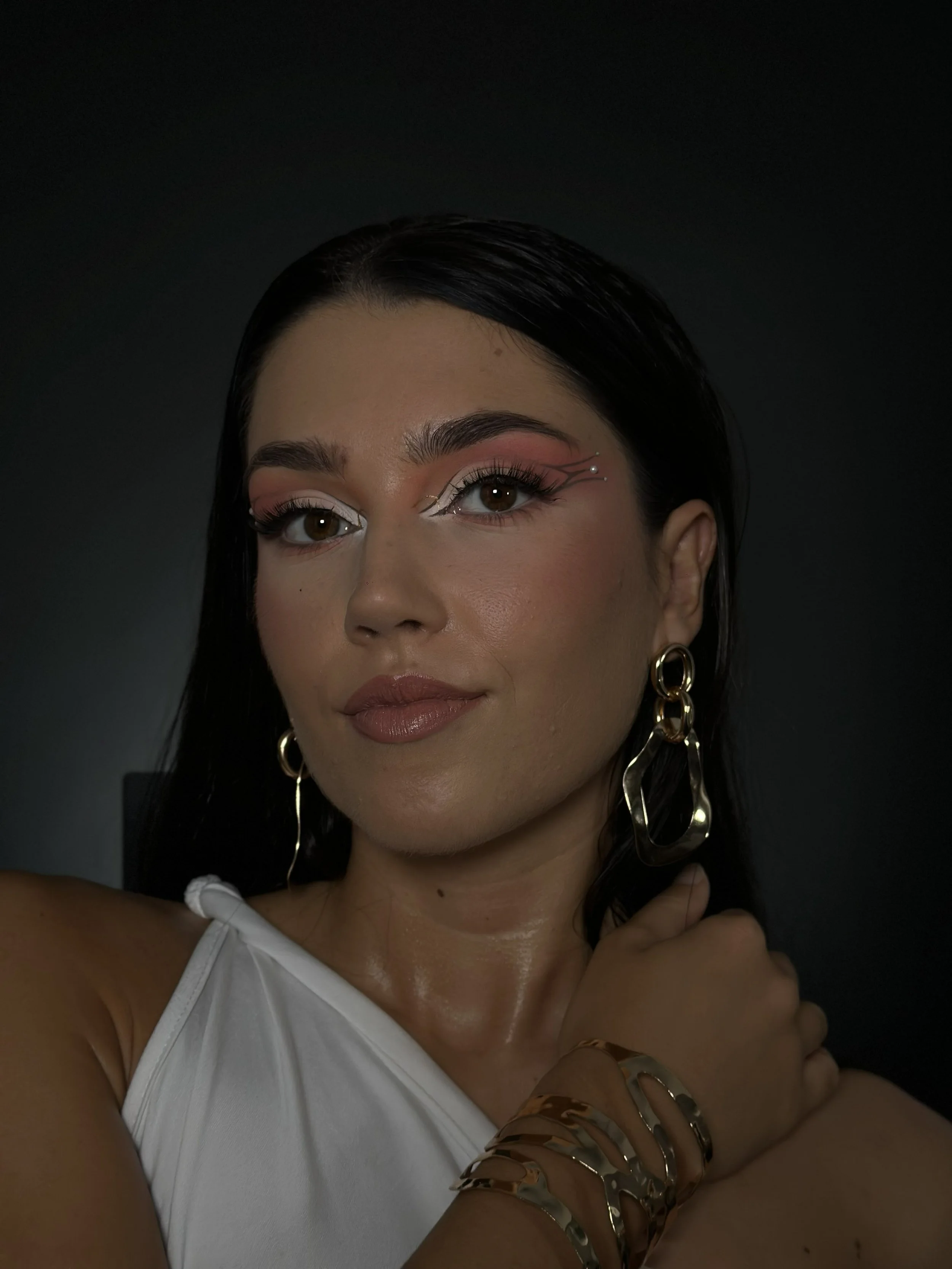 Close-up of a woman's face with dark hair, brown eyes, and makeup. She is wearing large gold earrings and multiple gold rings. She has a white top with thin straps.