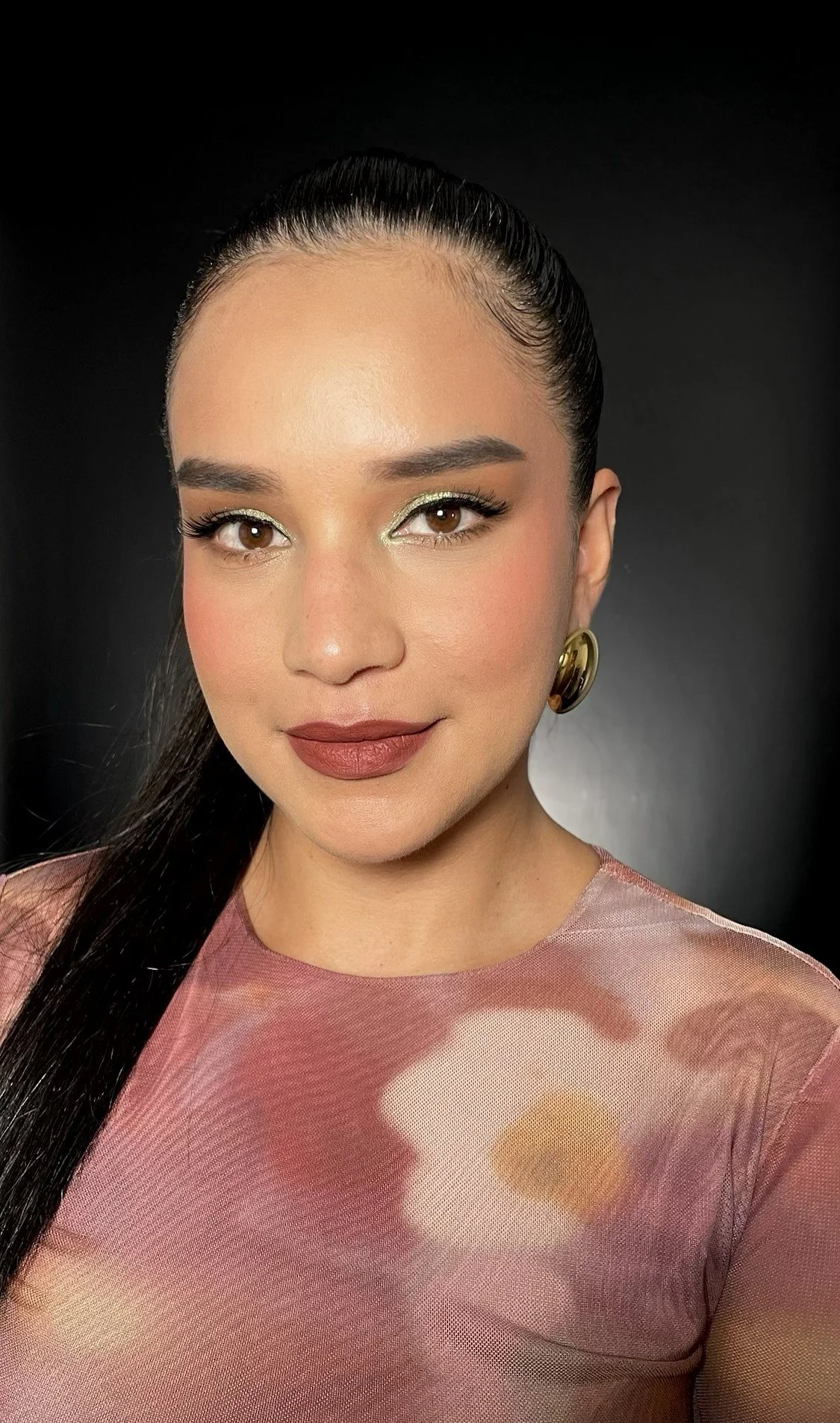 A woman with long dark hair pulled back, wearing gold earrings, makeup with gold eyeshadow, and a mauve top with sheer fabric, looking at the camera against a dark background.