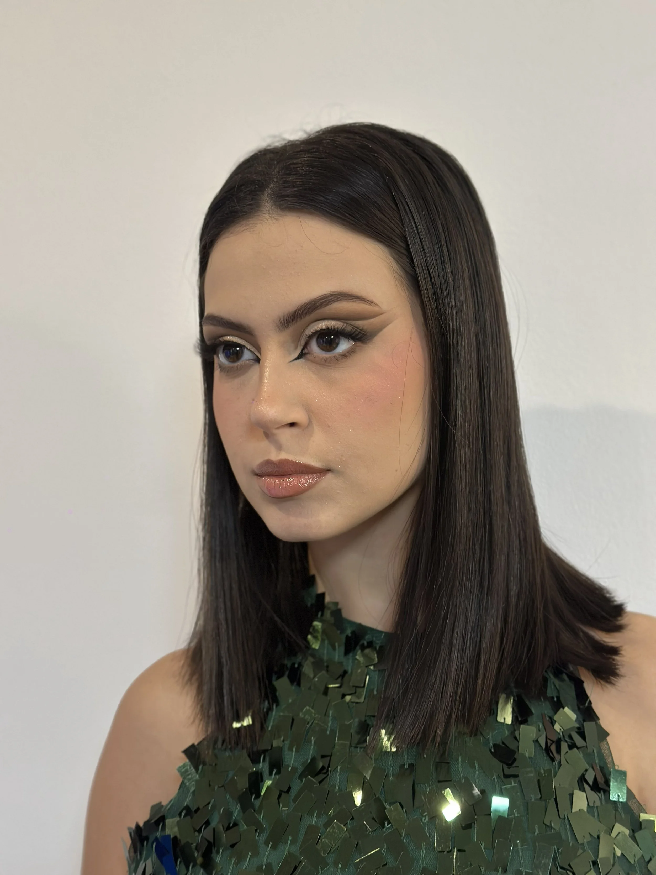 A woman with long dark hair and makeup, wearing a dress with reflective square sequins, standing against a plain white wall.