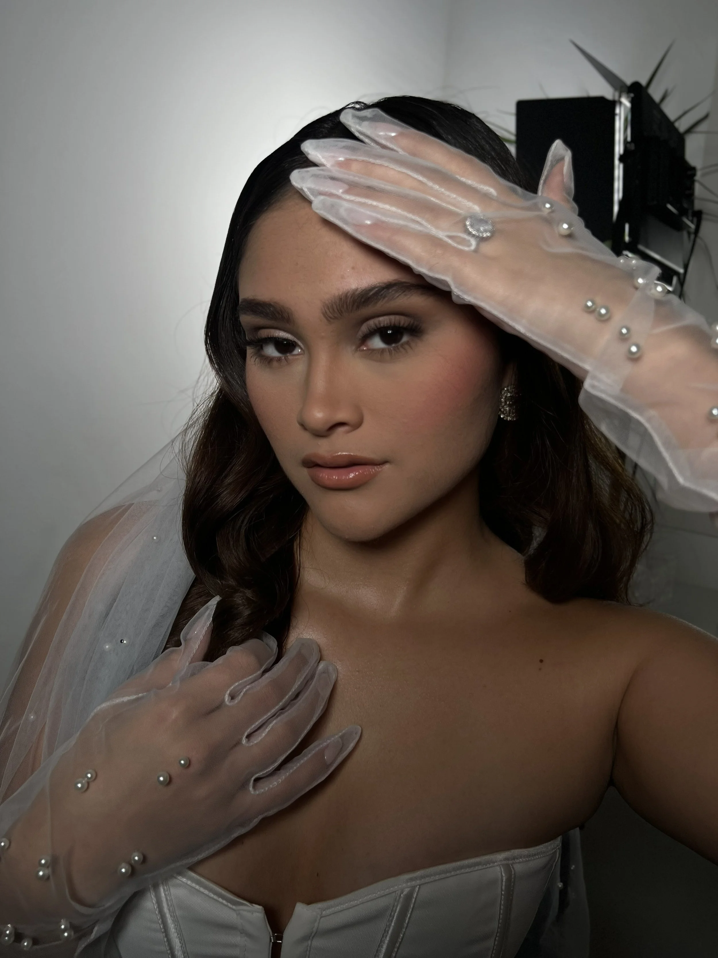 A young woman with dark hair and makeup posing with her hand on her forehead, wearing transparent gloves with pearl embellishments, a white corset, and earrings.