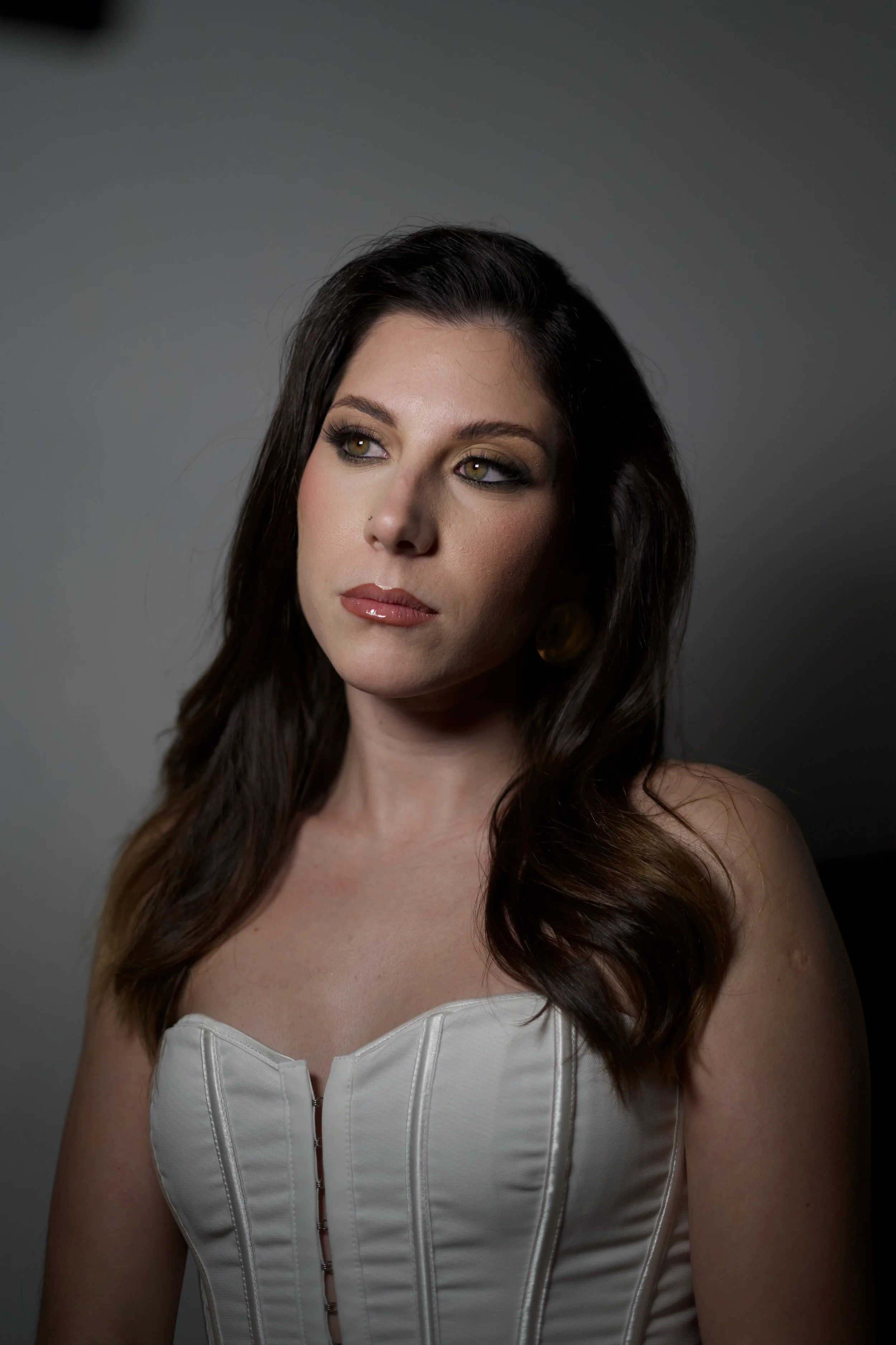 A woman with long brown hair and green eyes dressed in a strapless white dress with a front zipper, looking to her right against a gray background.