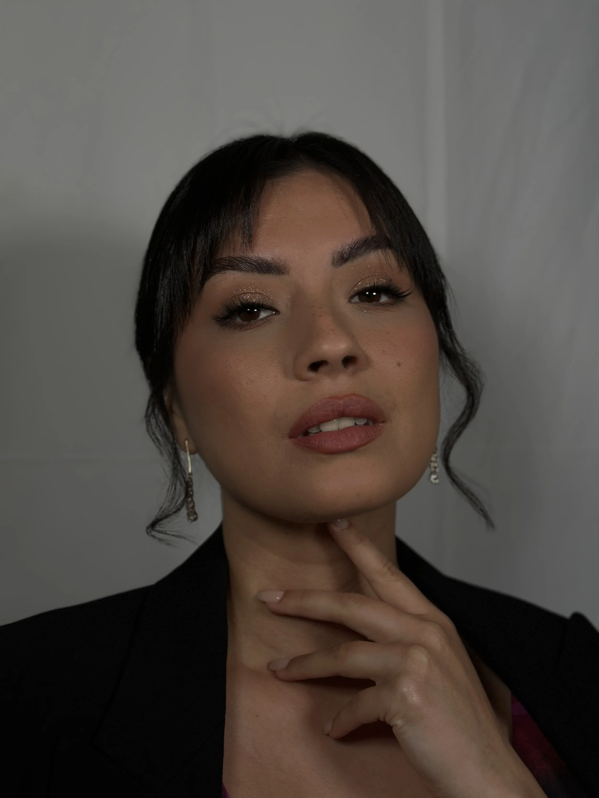 Close-up of a woman with short dark hair and makeup, touching her chin with her hand, wearing earrings and a black jacket.