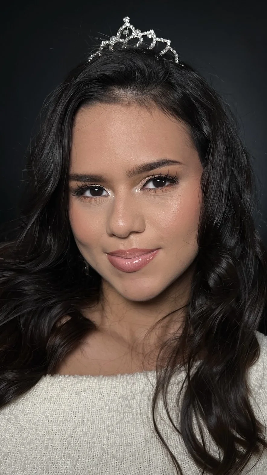 A young woman with long dark wavy hair wearing a small tiara, smiling softly, dressed in a light-colored top, with a dark background.