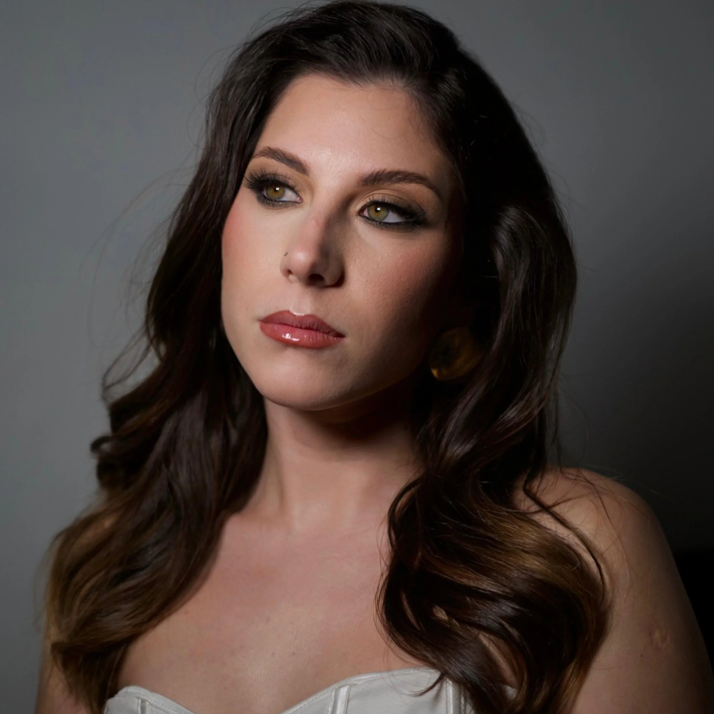 A woman with long dark hair and hazel eyes wearing a white top, looking to the side against a gray background.