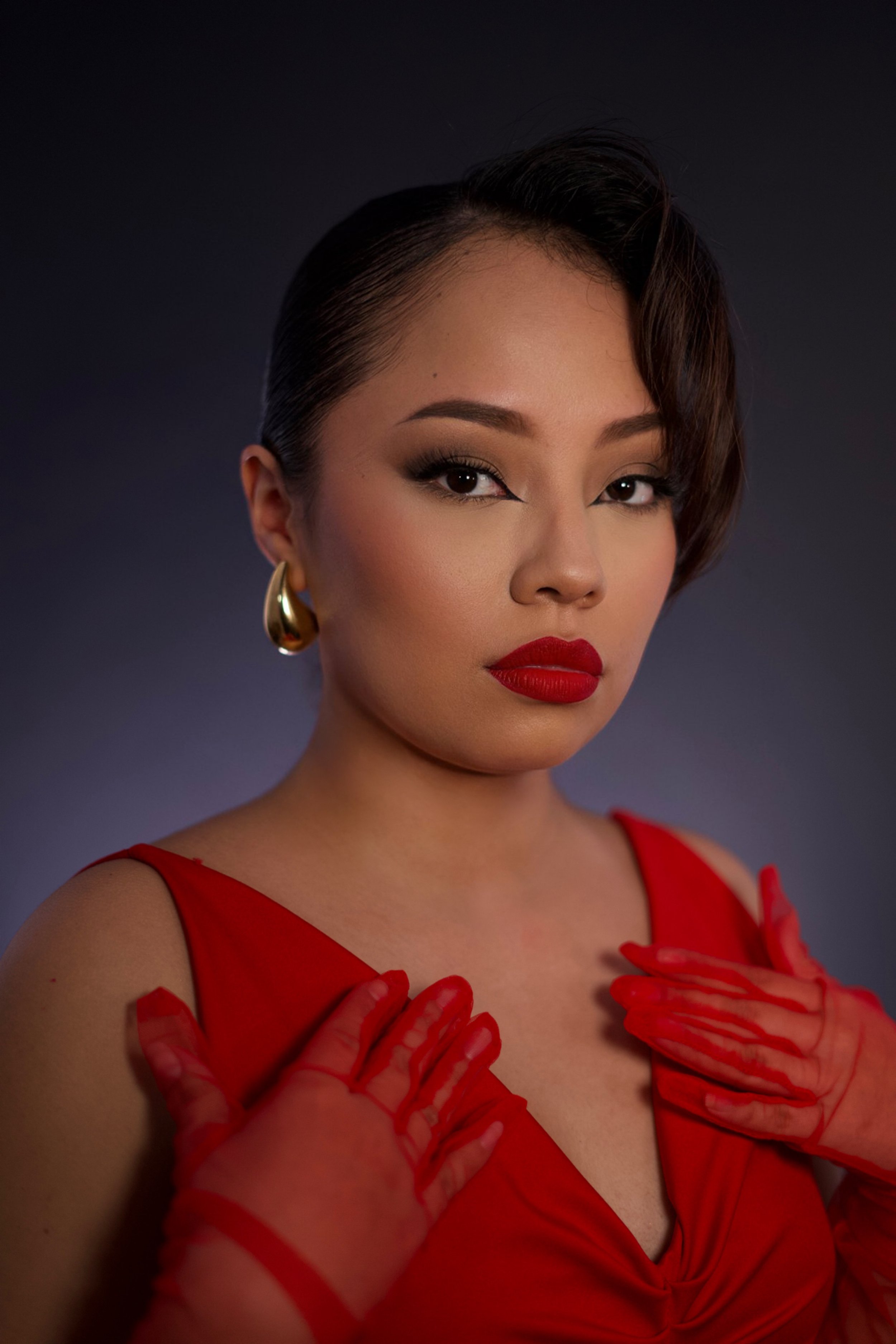 A woman with short dark hair styled to the side, wearing gold hoop earrings, red lipstick, and a red dress. She has makeup on and her hands, covered in red sheer gloves, are placed on her chest against a dark background.
