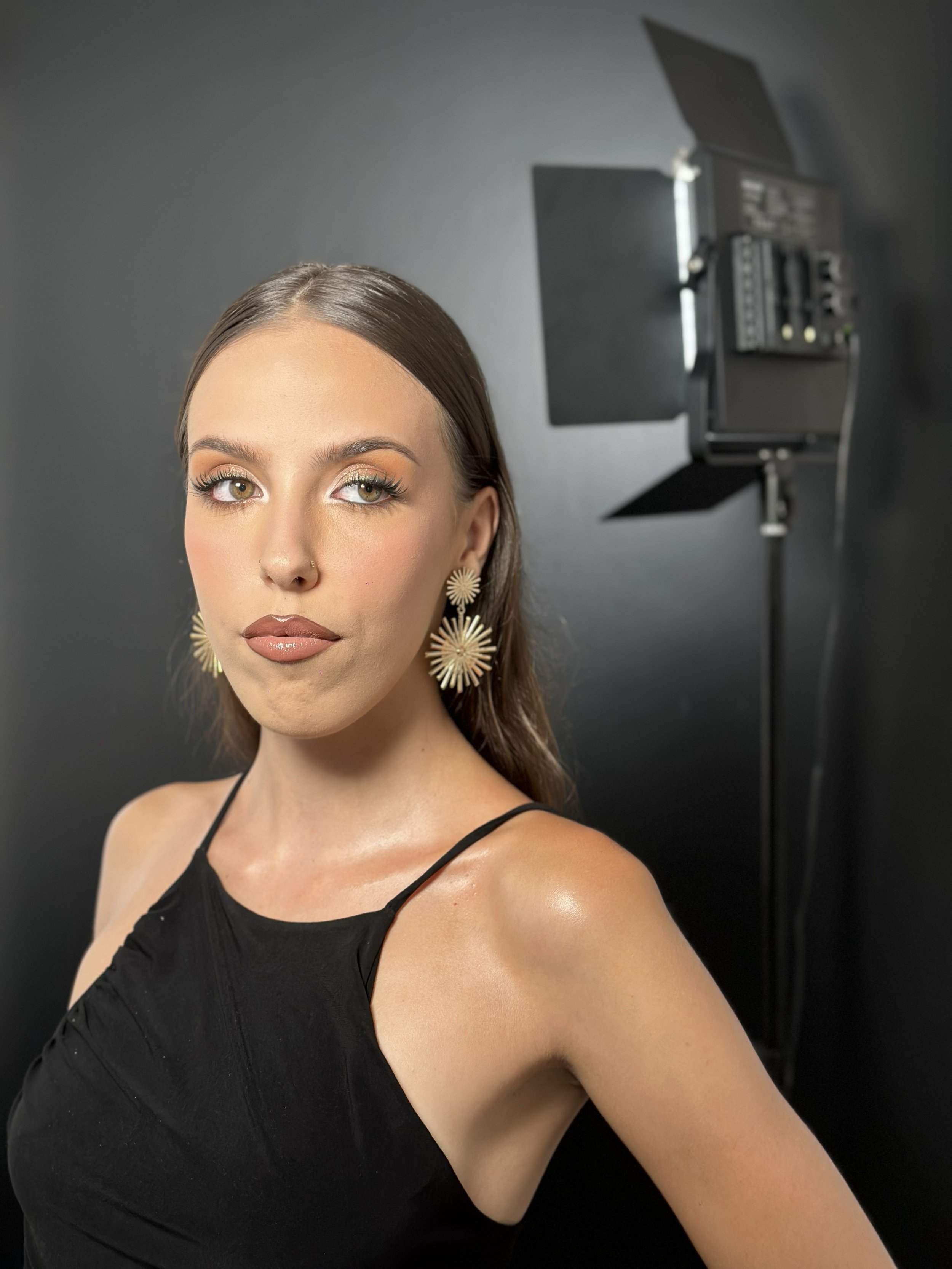 Close-up portrait of a woman with makeup and earrings, photographed in a studio with professional lighting equipment on a black background.