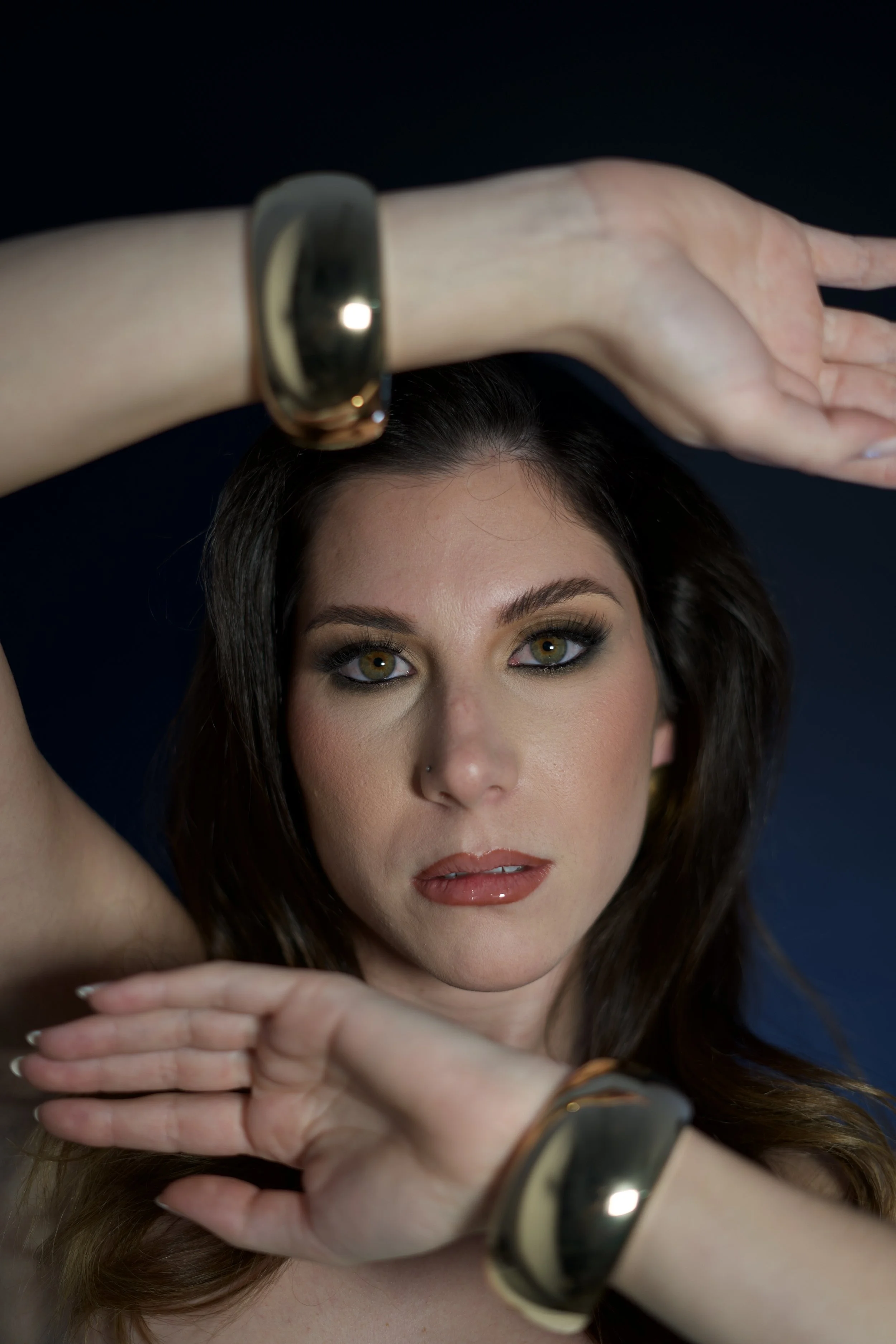A woman with long brown hair, makeup, and hazel eyes, is looking directly at the camera while raising her left arm over her head. She is wearing a large gold and black bracelet on her left wrist and a similar bracelet on her right wrist. The background is dark.