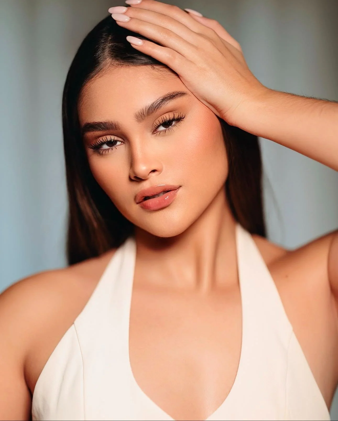 A woman with long dark hair, smooth skin, and wearing a white halter top, poses with her left hand on her forehead, looking directly at the camera.