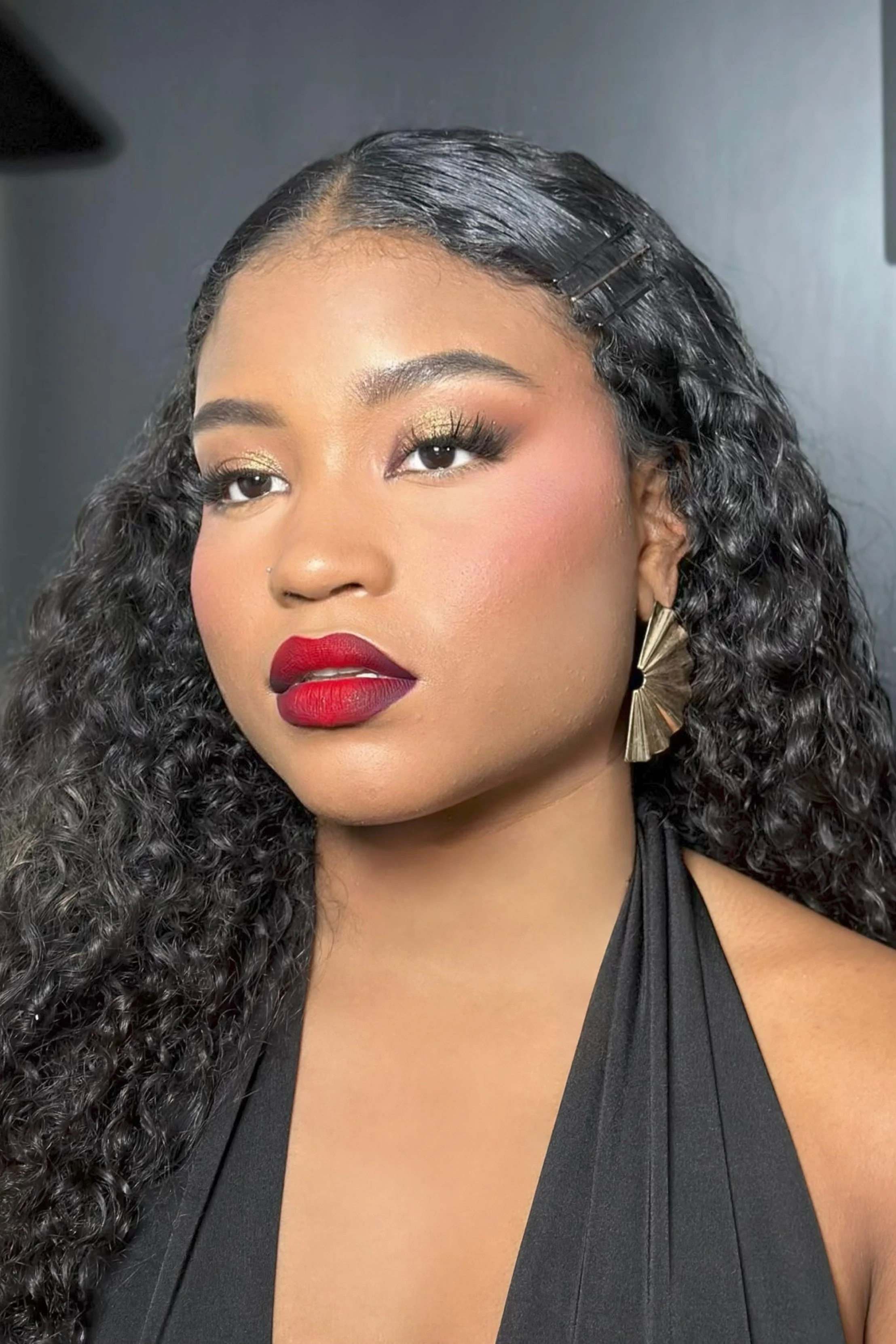 Close-up of a woman with dark curly hair, wearing gold earrings, red lipstick, and makeup, dressed in a black sleeveless top.