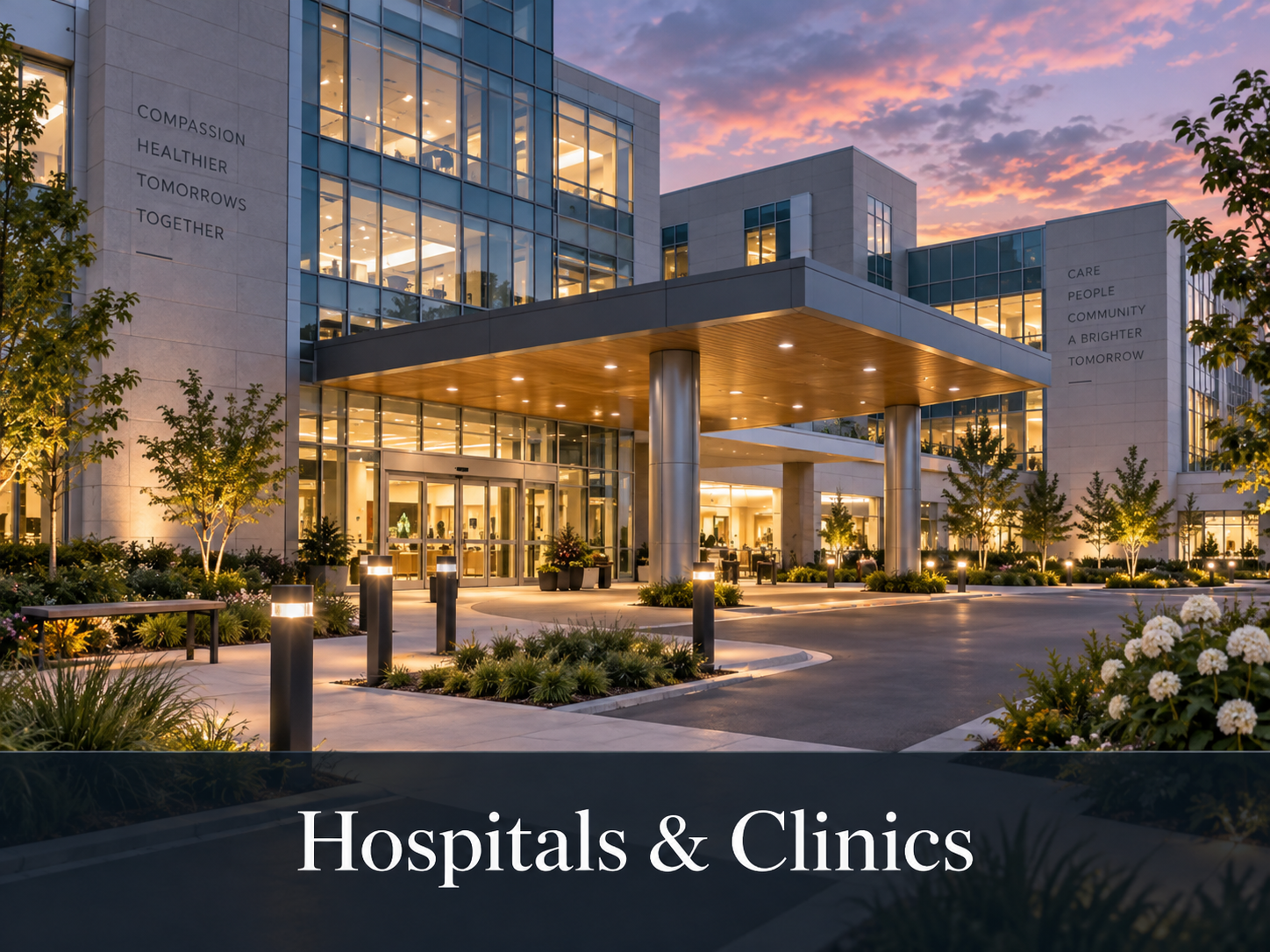 Exterior view of a modern hospital or clinic building at sunset, with illuminated windows, a landscaped entrance, and landscaping with trees and flowers.