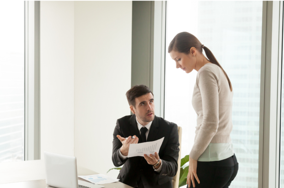 Businesswoman standing and talking to seated businessman in an office with large windows.