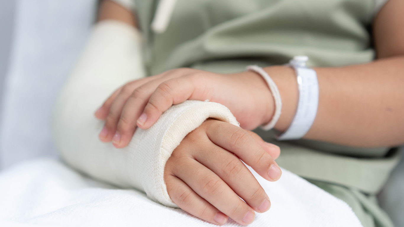 Close-up of a child's hands, one with a cast and the other with an IV wristband, resting on a hospital bed.