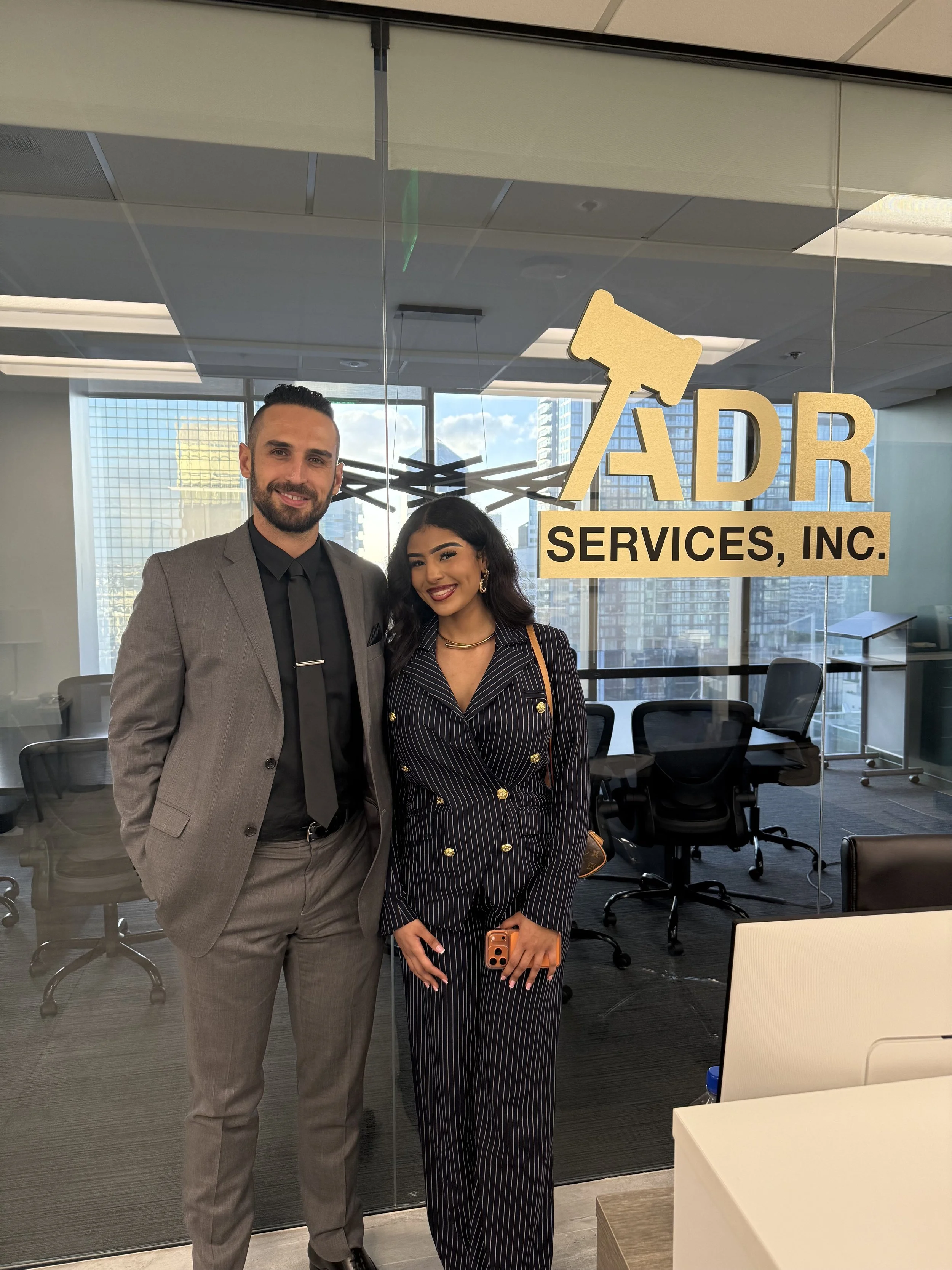 A man in a gray suit and a woman in a striped suit with gold buttons and jewelry are standing together in an office, smiling. Behind them is a logo for 'ADR Services, Inc.' with a gavel icon, and large windows showing a cityscape.