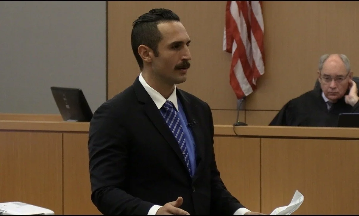 A man with dark hair, a mustache, and wearing a suit and tie stands in a courtroom, holding some papers. In the background, a judge sits at a bench, wearing robes, with an American flag behind him.
