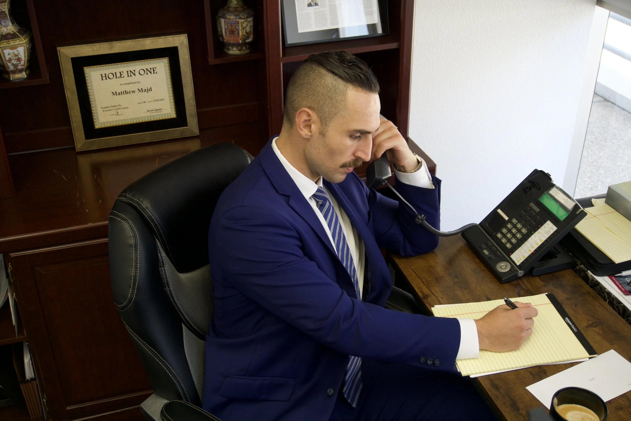 A man wearing a blue suit with a striped tie sitting at a wooden desk, talking on a corded phone, with a notepad and pen in hand, in an office with wooden shelves and a framed certificate on the wall.