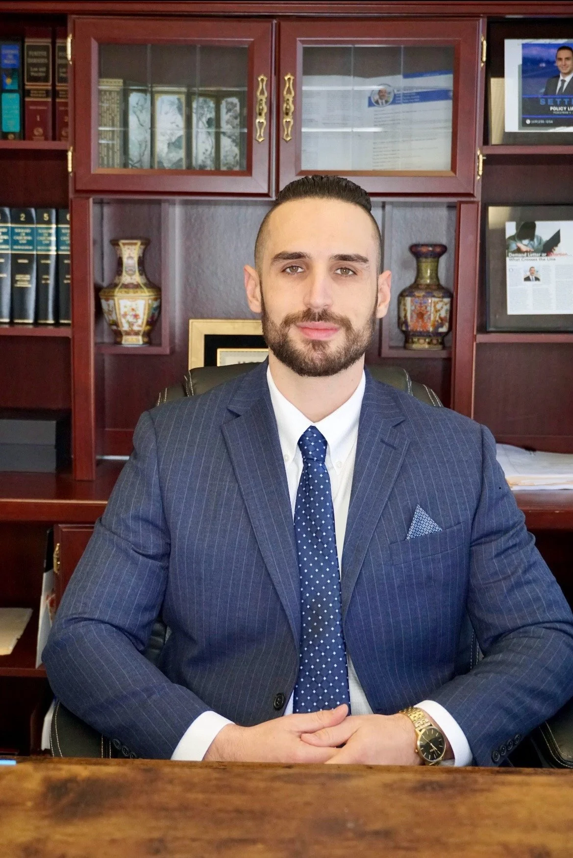 A man in a dark blue pinstripe suit with a white shirt and a blue polka dot tie sitting at a wooden desk in an office. He has short dark hair, a beard, and is wearing a gold watch. Behind him are dark wood bookshelves with decorative vases and framed documents, and a flat-screen monitor showing a news broadcast.