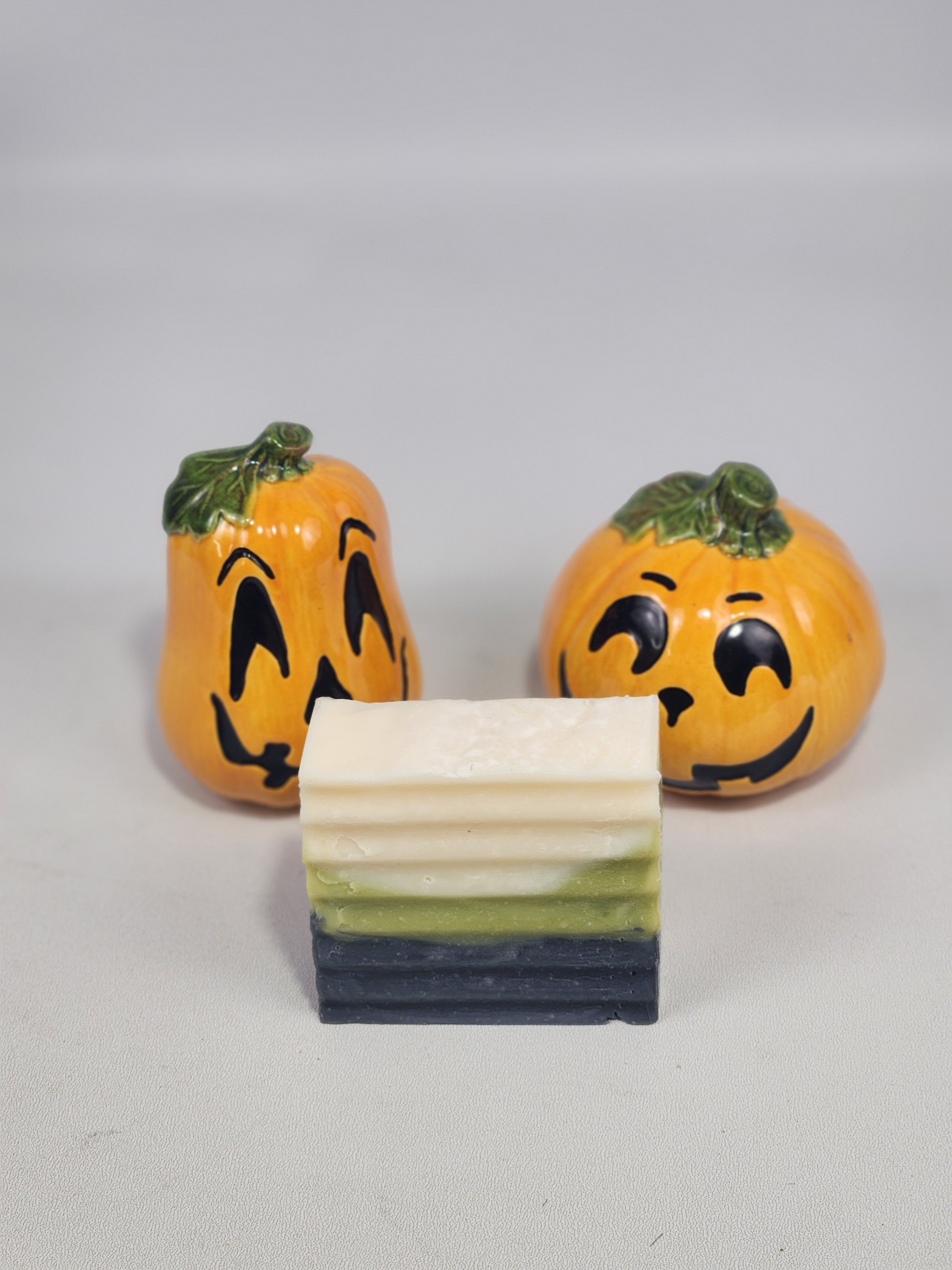 Two ceramic pumpkin-shaped figurines with painted faces, resembling jack-o'-lanterns, placed behind a white soap bar on a gray surface, against a plain gray background.
