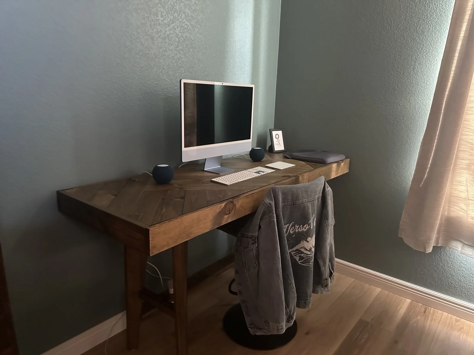 A wooden desk with a computer monitor, keyboard, and mouse, against a green wall. There are two small speakers, a framed picture, a closed laptop, and a jacket hanging on a chair, with a window and curtains in the background.
