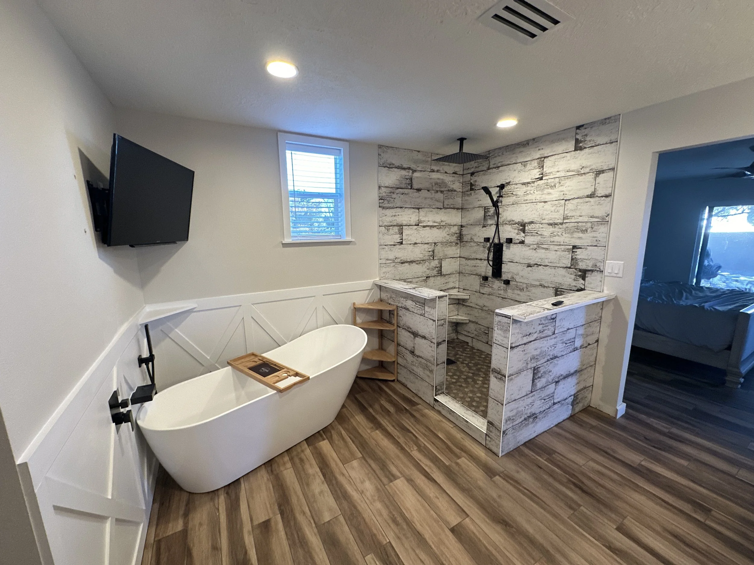 A modern bathroom with a white freestanding bathtub, a wooden stool, and a walk-in shower with a rustic wood tile wall. There is a window with white blinds, a mounted flat-screen TV, and recessed ceiling lights.