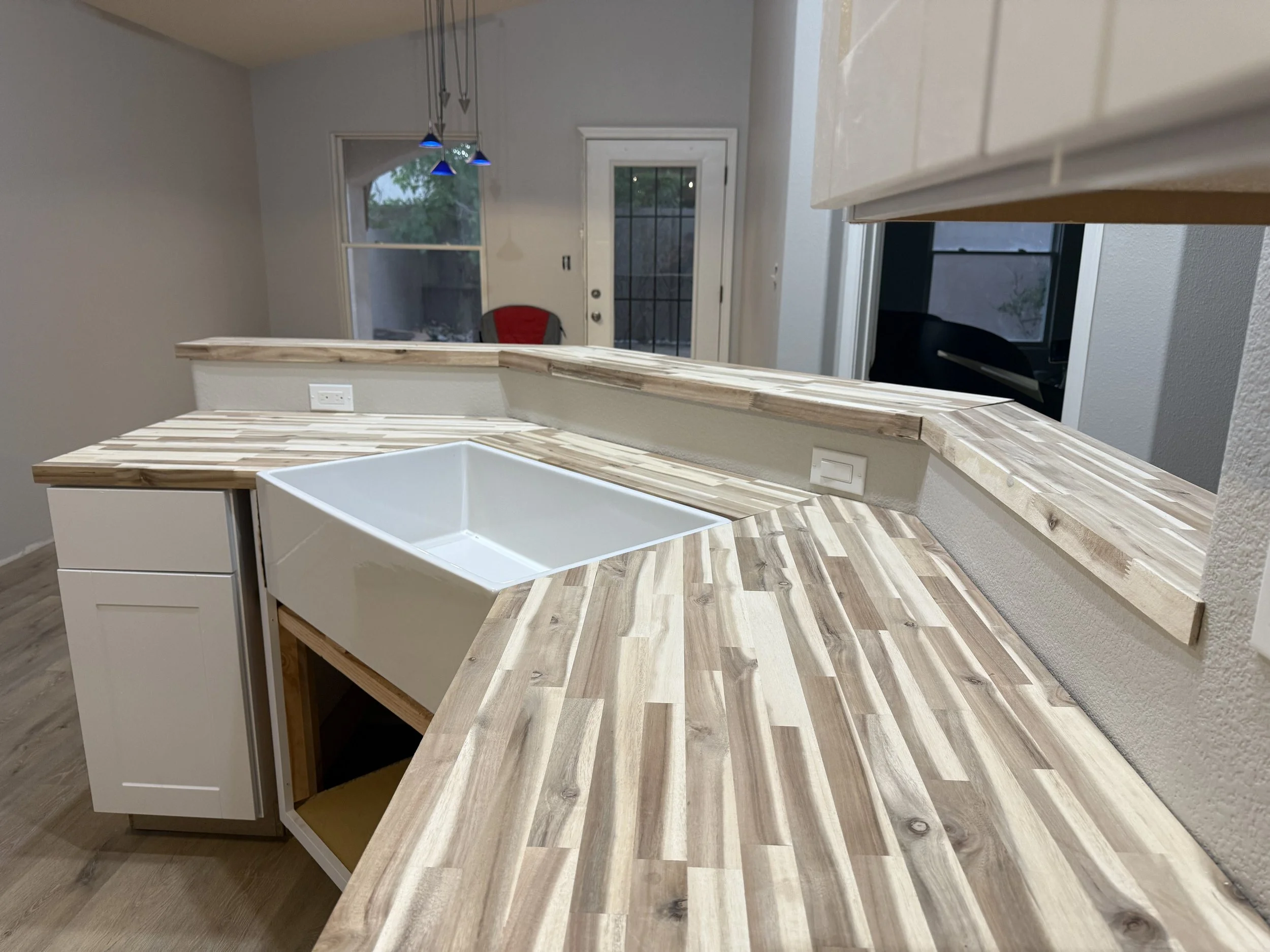 Partially completed kitchen interior with a wooden countertop, a white farmhouse sink, and a background view of sliding glass door and windows.