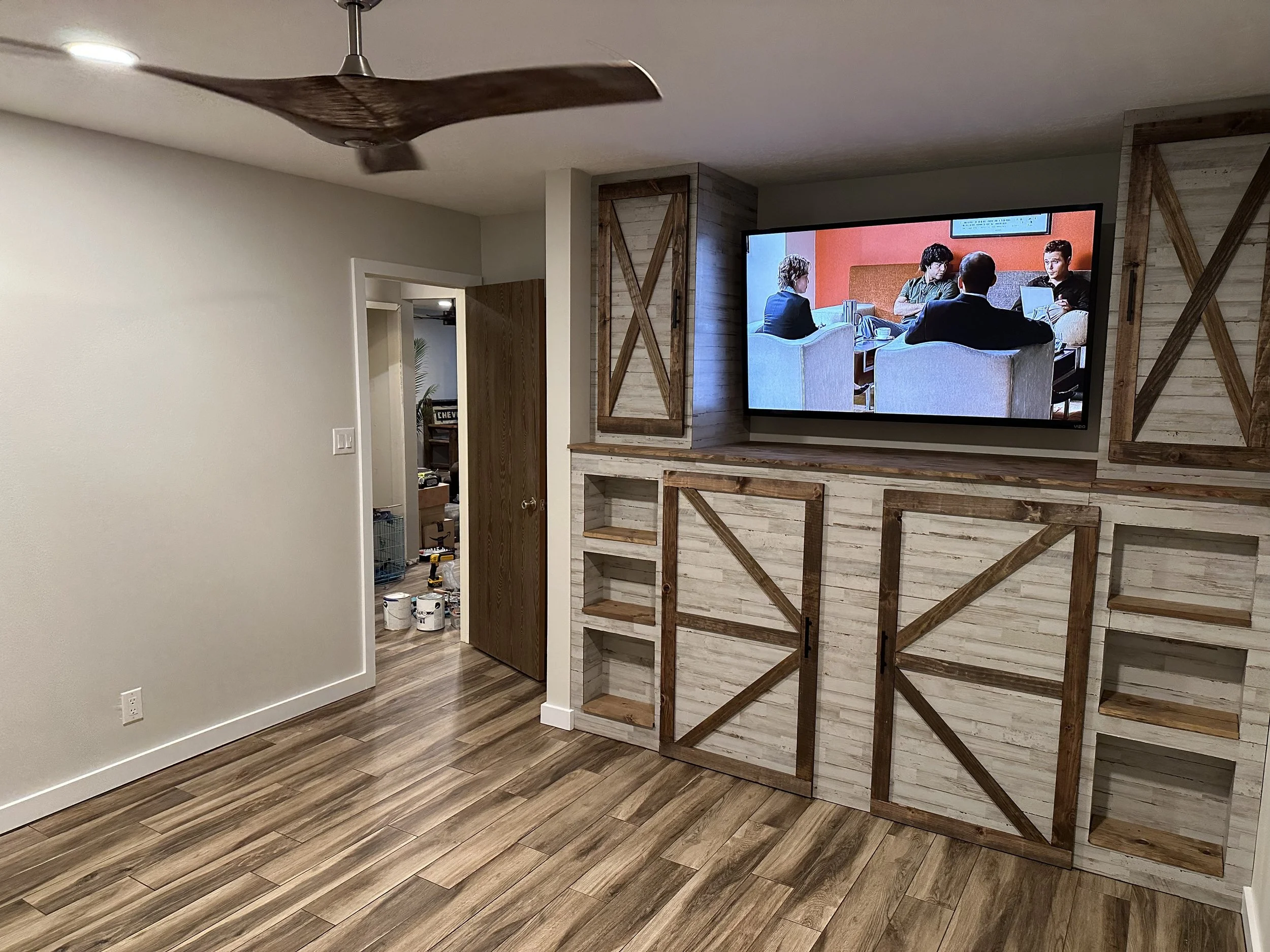 Bedroom with a mounted TV showing a video call with four people, a ceiling fan, wooden shelving, white walls, and hardwood floors.