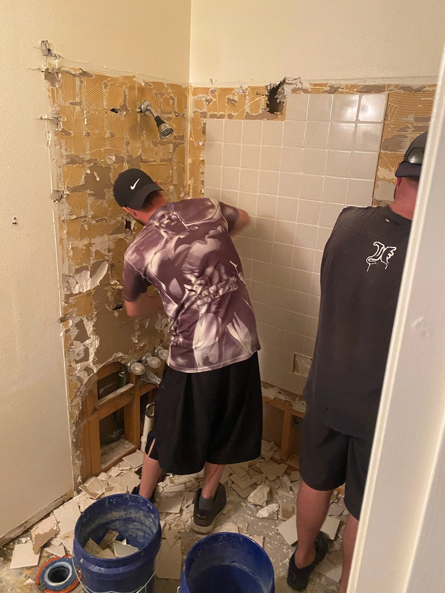 Two people working on a bathroom renovation. One person is breaking or removing tiles from the wall, while the other observes. Tiles and debris are scattered on the floor, and buckets are nearby.