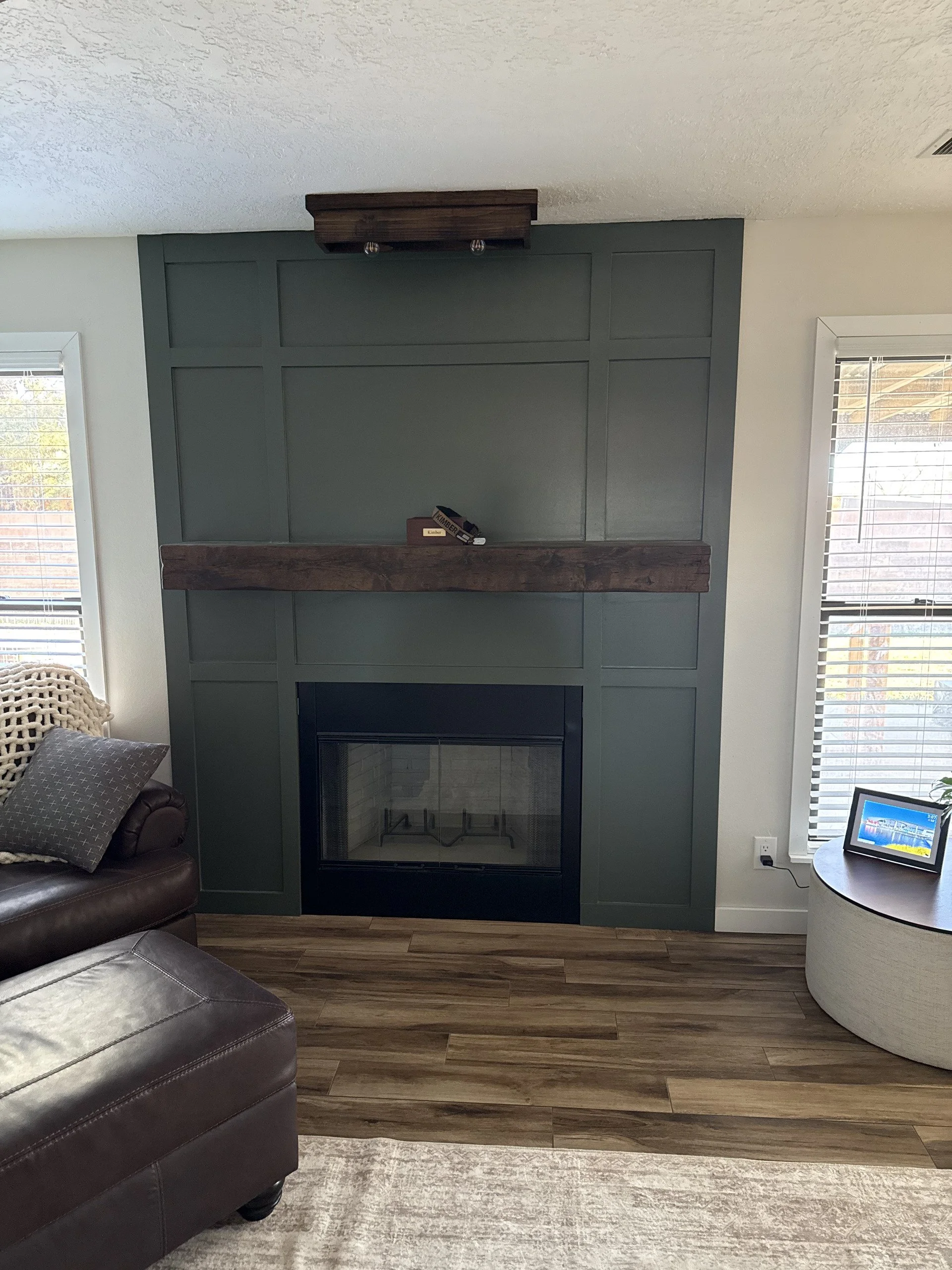 Living room with a green fireplace wall, wooden mantel, and two windows with white blinds. There is a brown leather chair with a cushion and a woven blanket, a small side table with a digital picture frame, and a beige rug.