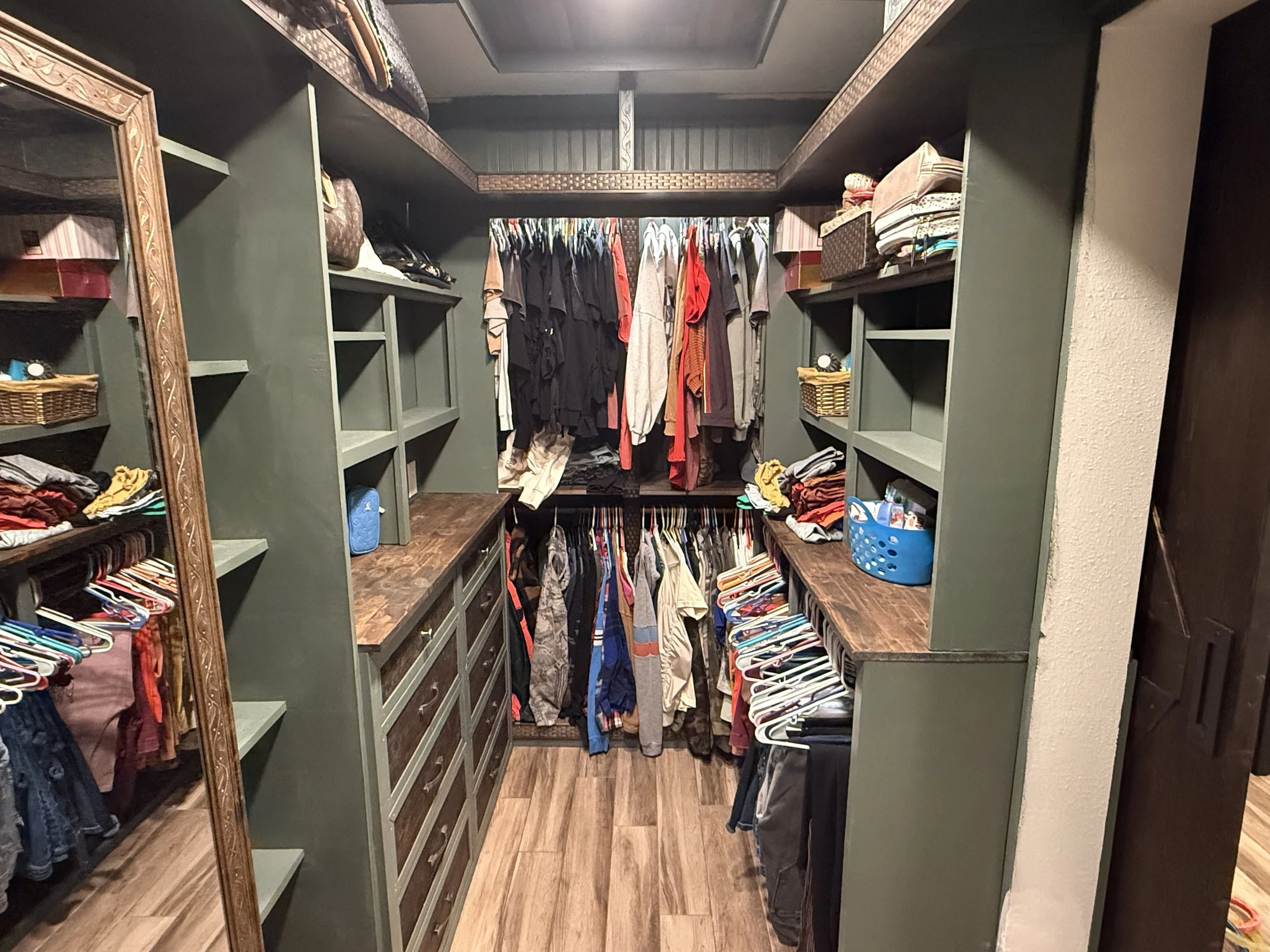 A walk-in closet with wooden flooring and dark gray walls. It features built-in green shelves on both sides filled with clothes, towels, and storage boxes. A rack with hanging clothes is in the center, with shelves below containing folded clothes and