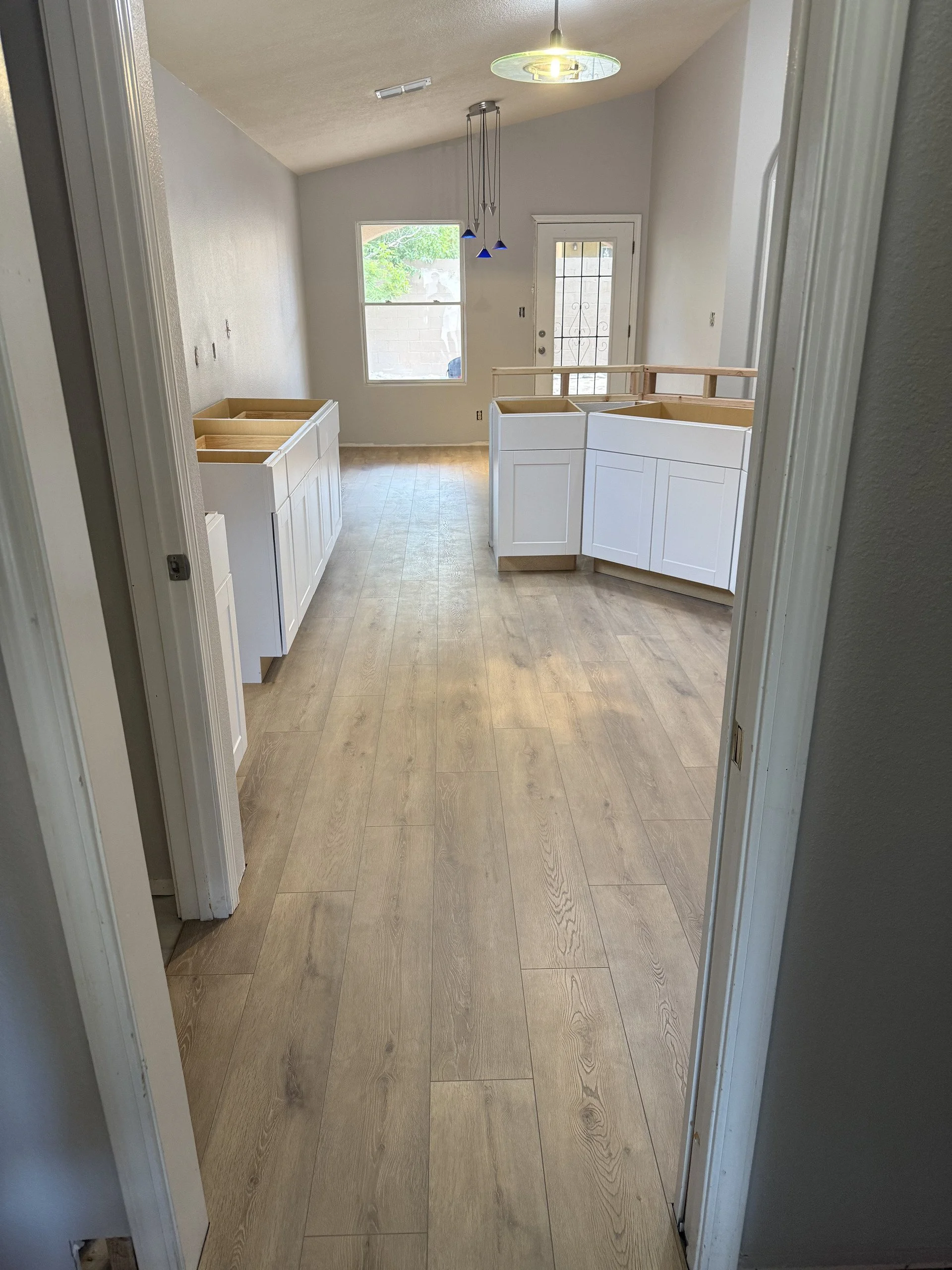 Empty kitchen space with light wood flooring, white cabinets without countertops, a window, and a door with a decorative metal grille.