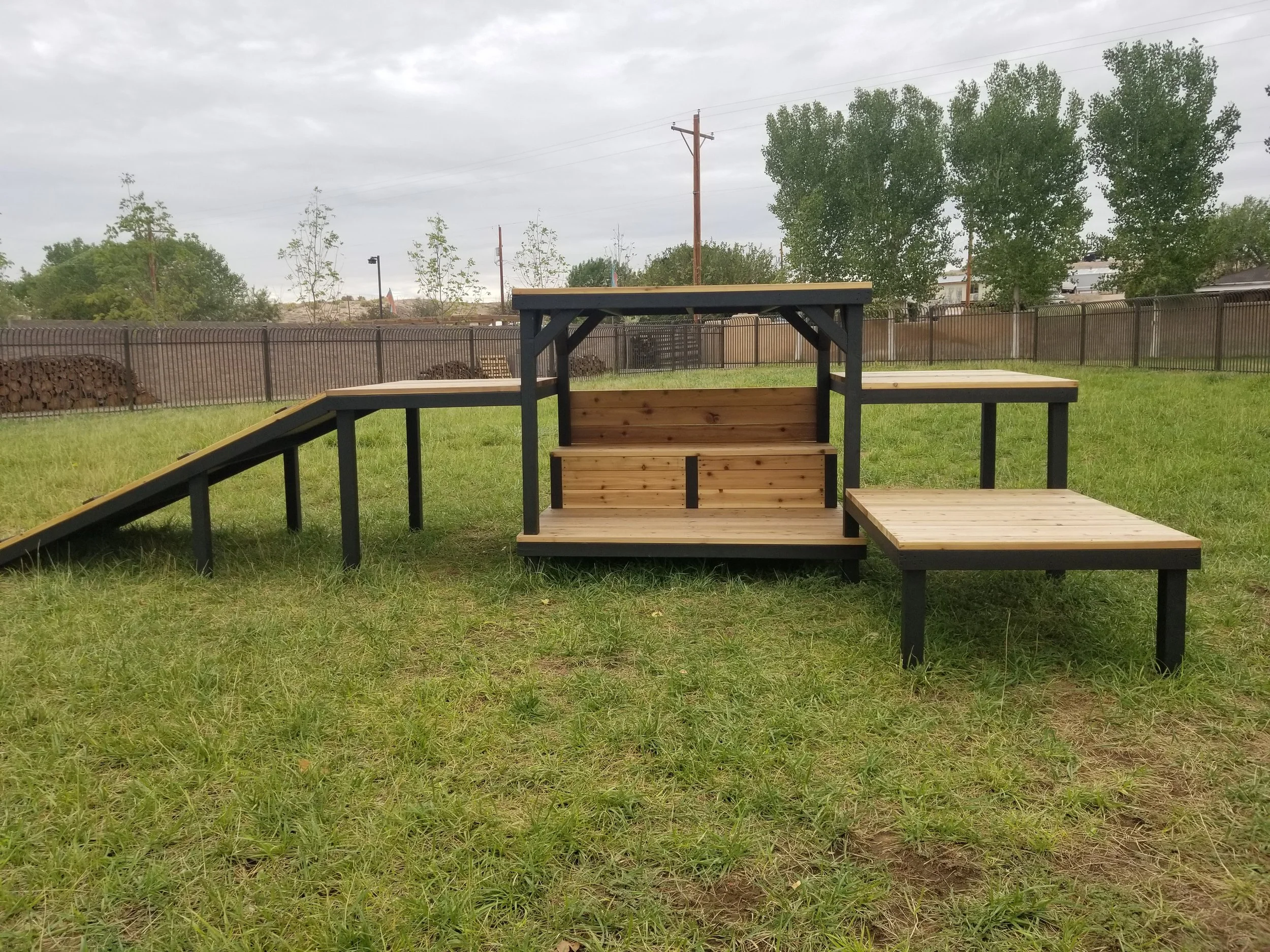 A backyard wooden structure, green grass, trees, a fence, and cloudy sky.