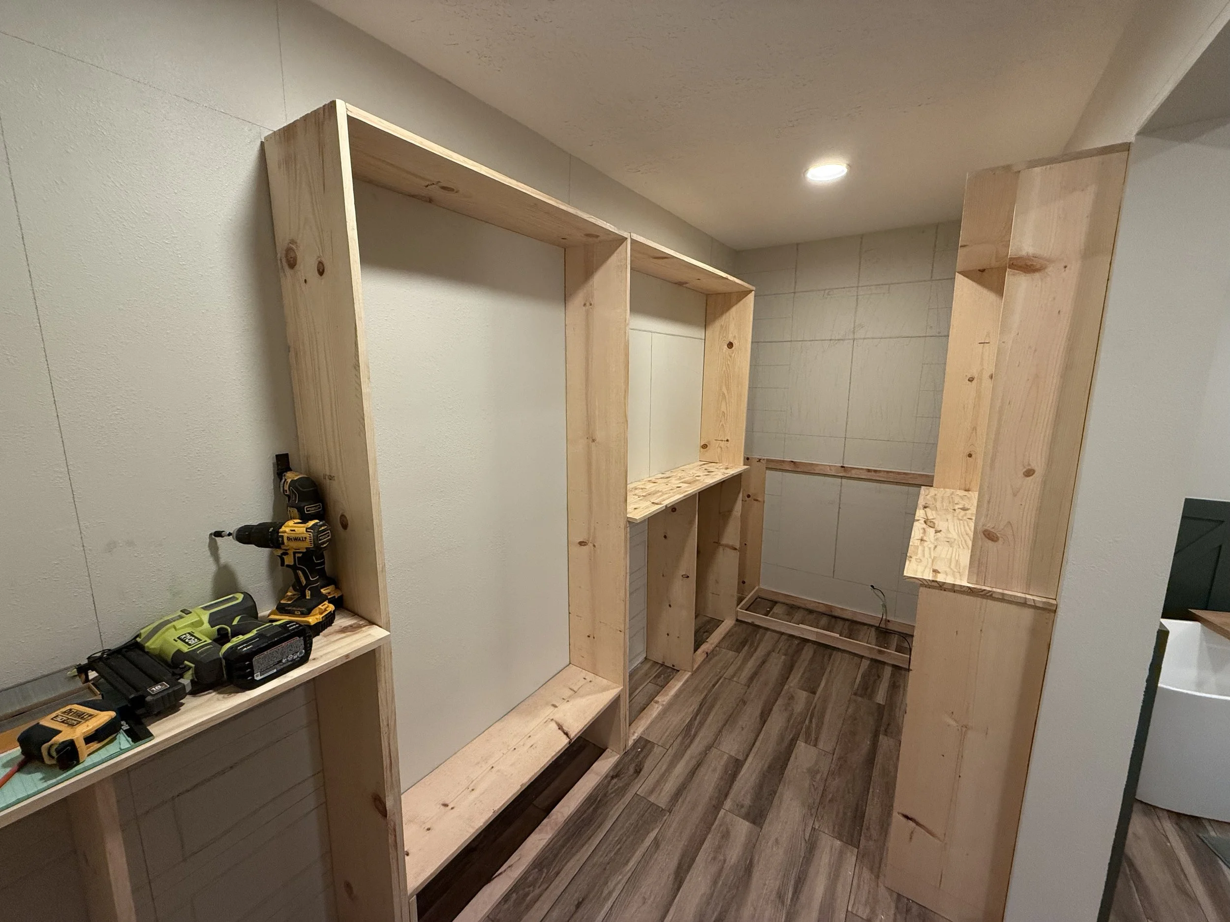 Unfinished wooden shelving units being assembled on a wall in a room, with tools on a nearby table.