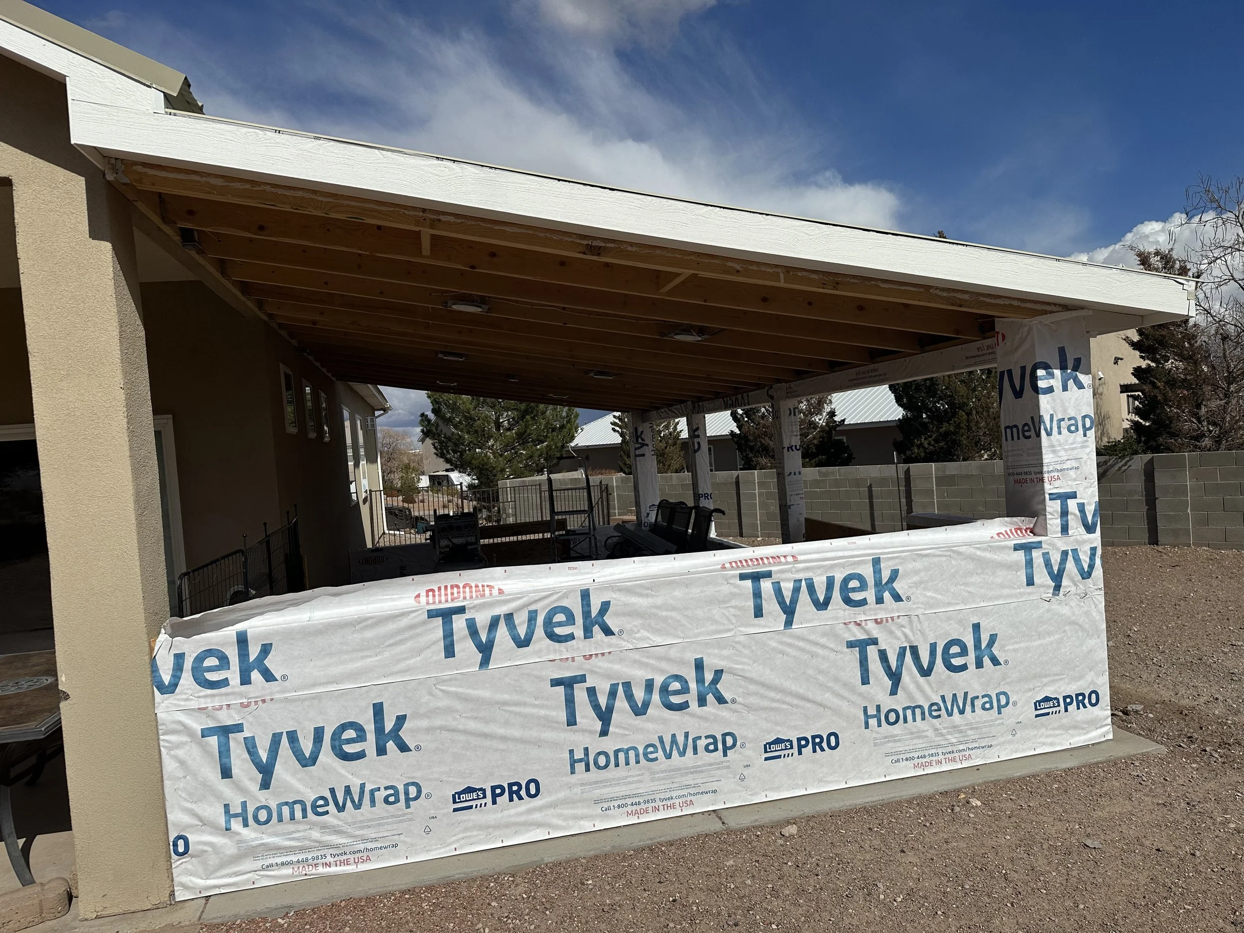 Construction site with house exterior, showing a partially built porch with white Tyvek wrap, wooden beams, and a beige stucco wall. The sky is partly cloudy with blue sky visible.