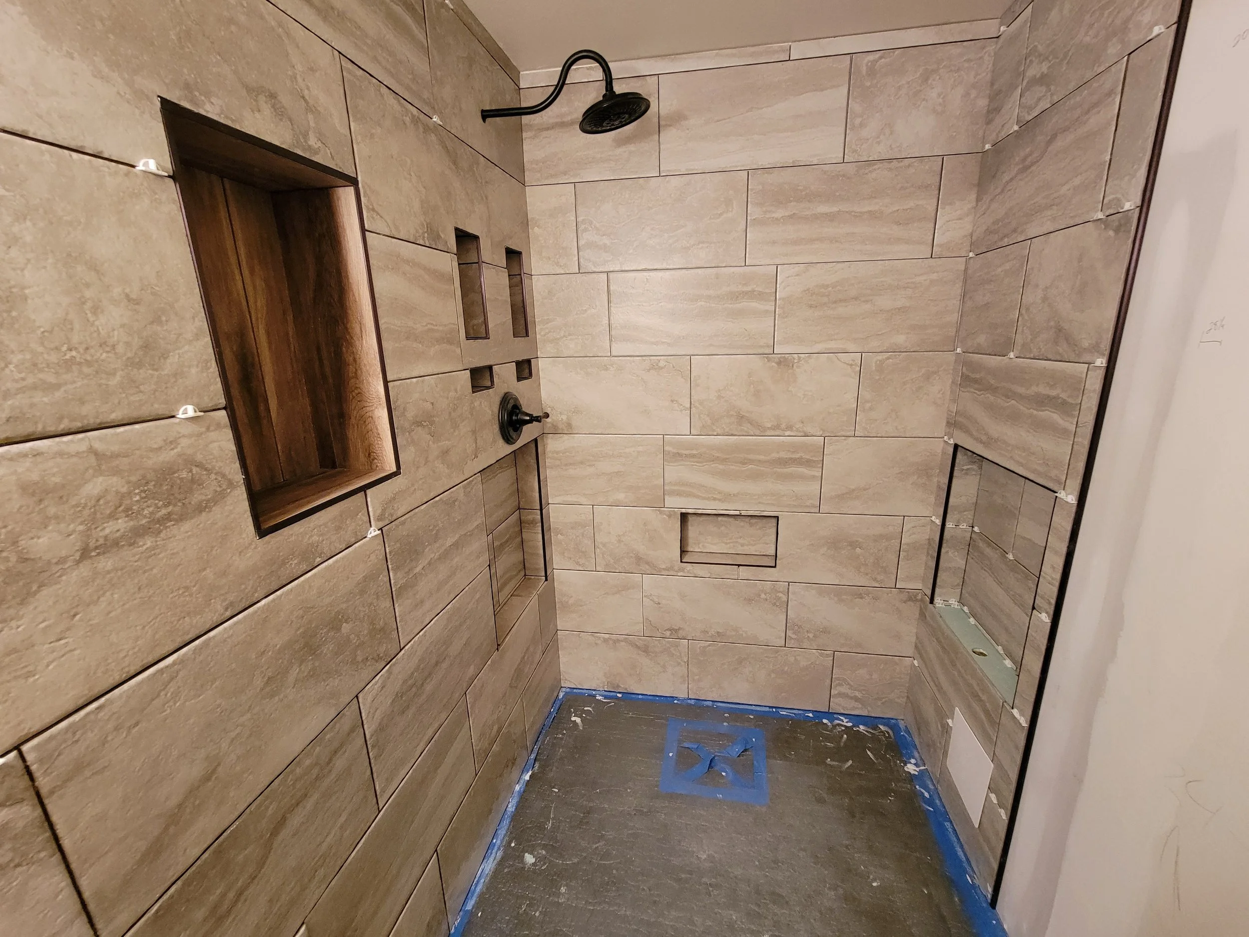 A tiled shower with beige tiles, a dark showerhead, built-in niches, and a partially finished wall with construction supplies and drywall.