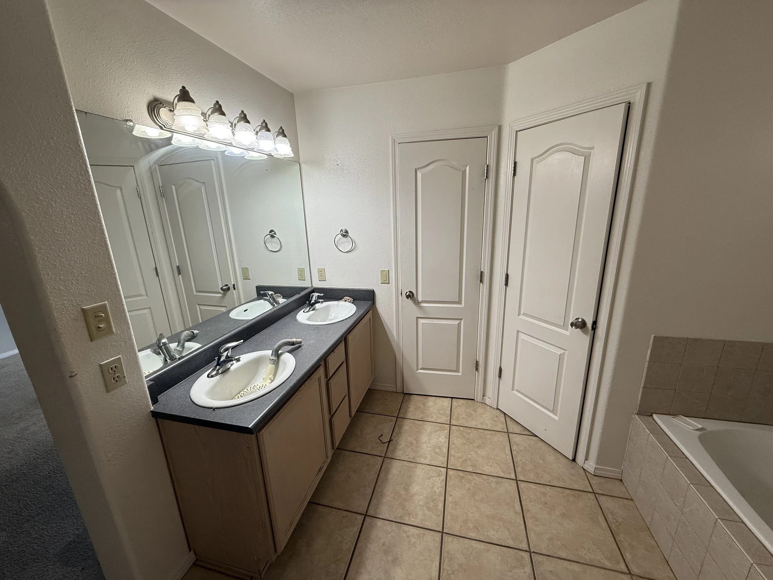 A bathroom with dual sinks on a dark gray countertop, two mirrors, light fixtures with multiple bulbs above the mirror, white cabinets below the sinks, tiled floor, and closed white doors.