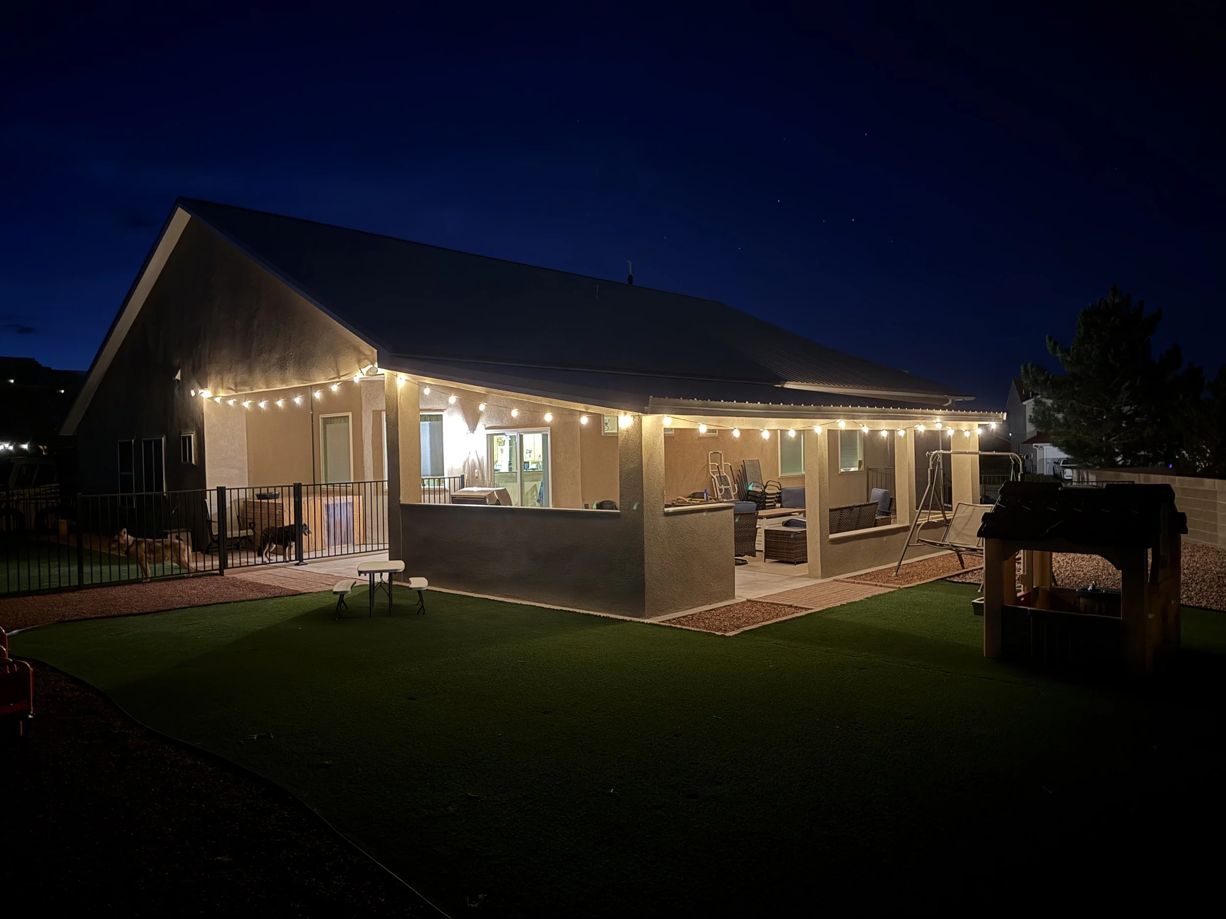 Covered patio illuminated by string lights. The house has a steep roof, a patio with furniture, and a backyard with a swing set, a dog, and a grassy area. The sky is dark with visible stars.